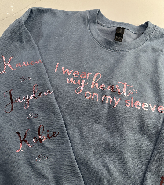 Light blue sweatshirt with embroidered names 'Kawen', 'Jaxson', and 'Kobie' on the sleeve and the phrase 'I wear my heart on my sleeve' embroidered on the chest.