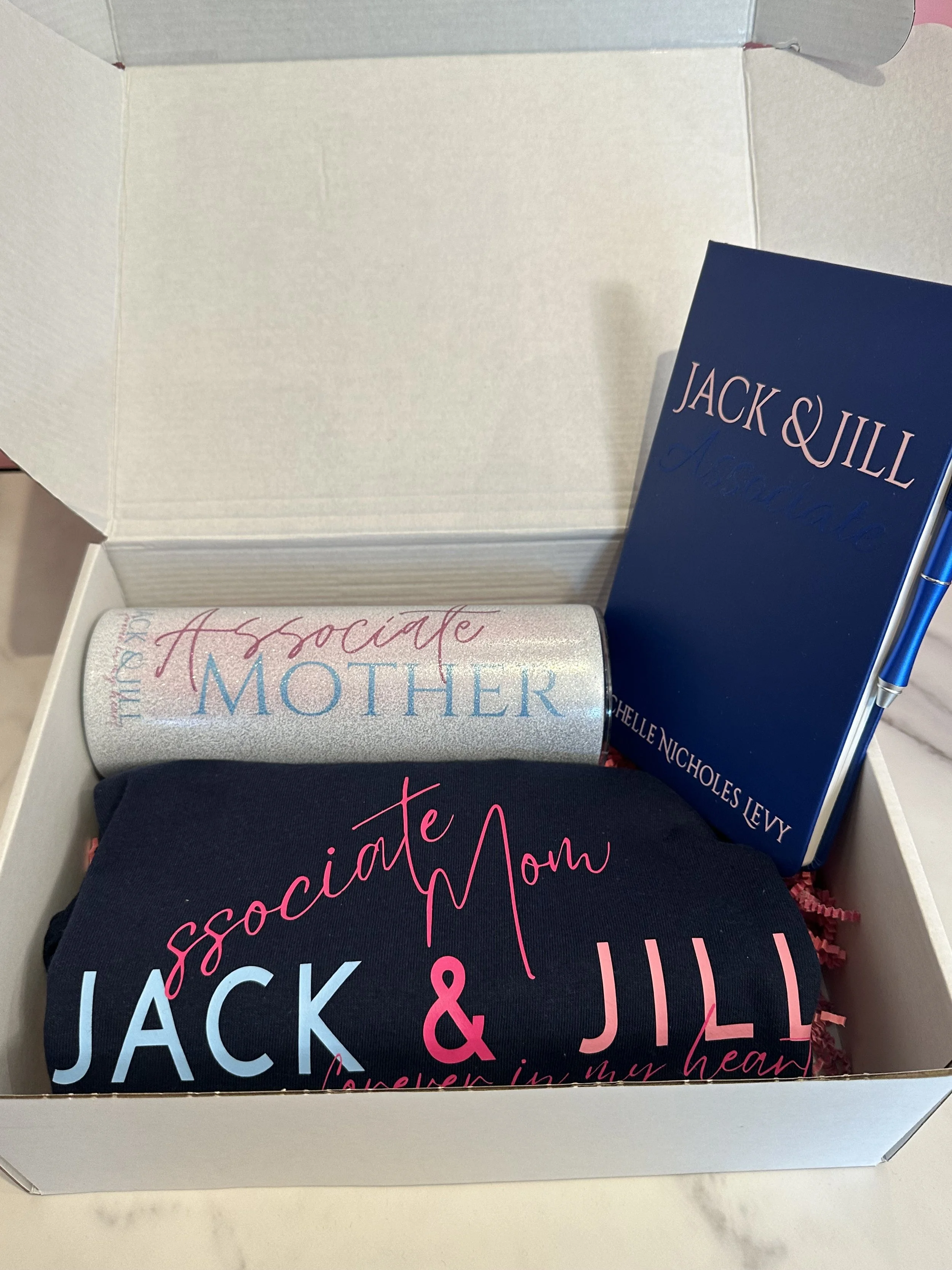 Open gift box containing a navy blue shirt with pink and light blue text, a blue notebook titled 'Jack & Jill' by Alehelle Nichols Levy, and a glittery silver tumbler with pink and blue text.