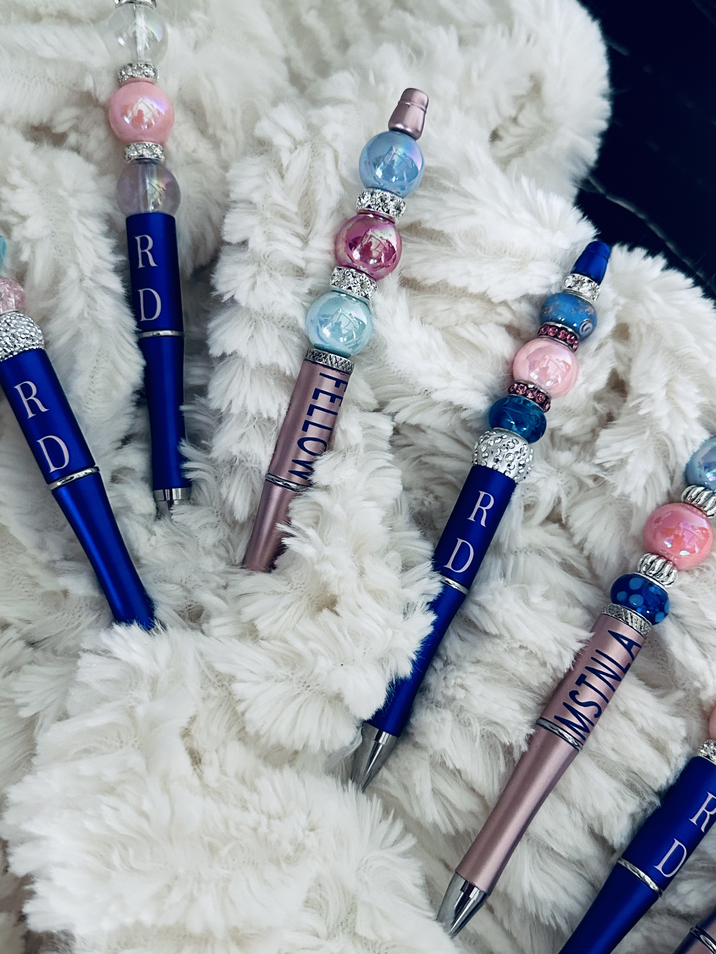 Close-up photo of decorated pens with pink, blue, and clear beads and rhinestones, resting on a soft, fluffy white fabric.