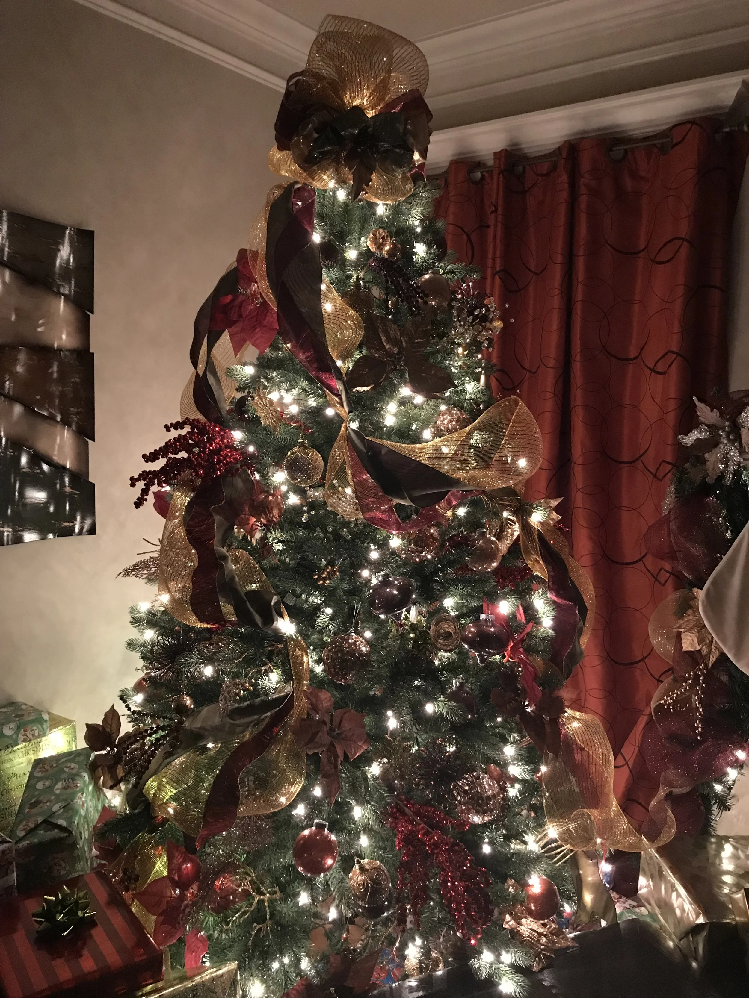 Decorated Christmas tree with gold and burgundy ribbons, ornaments, and lights, surrounded by wrapped presents.