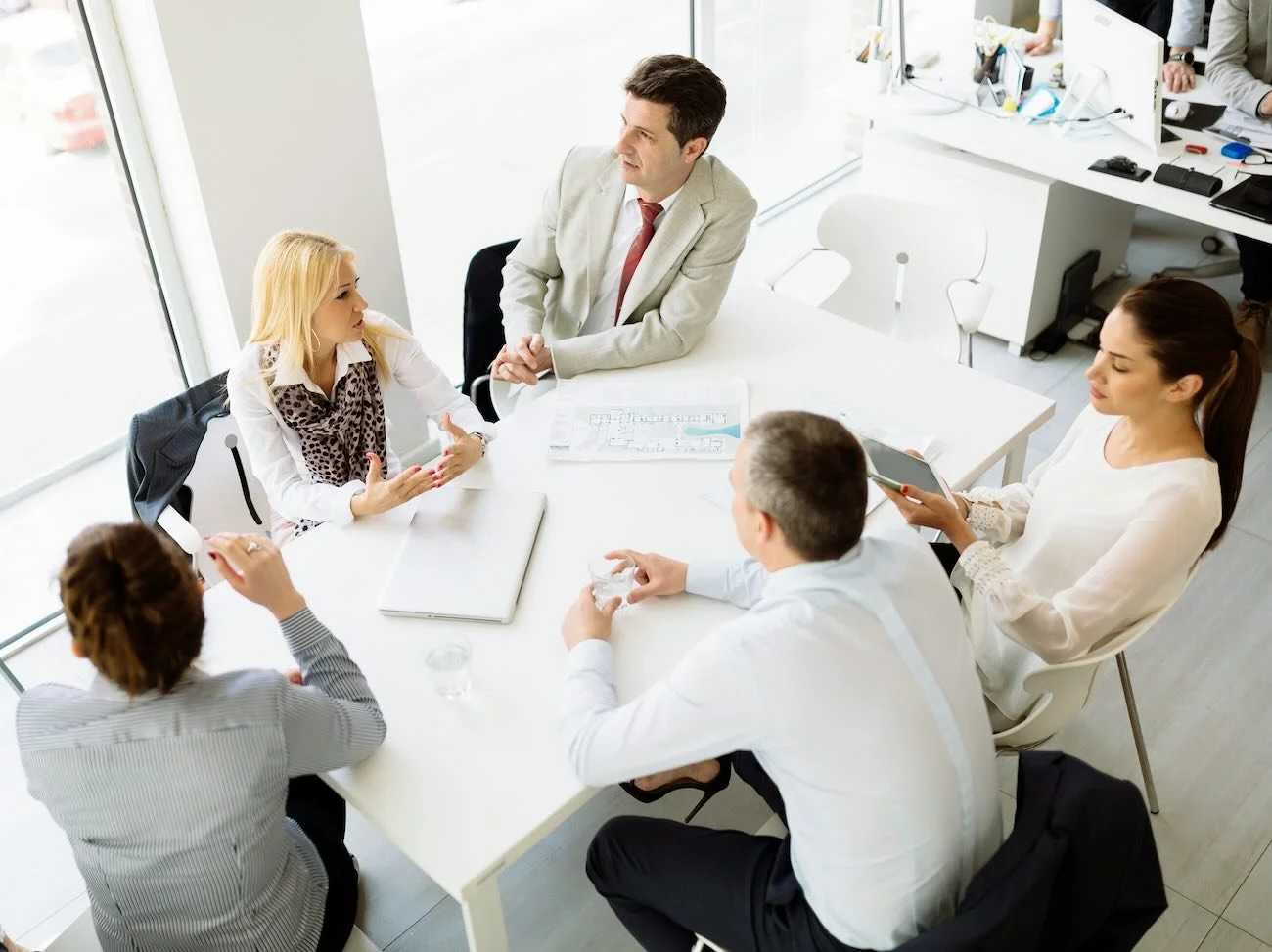 Business meeting with five people, three women and two men, sitting around a white table in a modern office, discussing and taking notes.