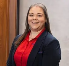 A woman with long brown hair smiling, wearing a navy blazer and red shirt, standing in front of a wooden door and gray wall.