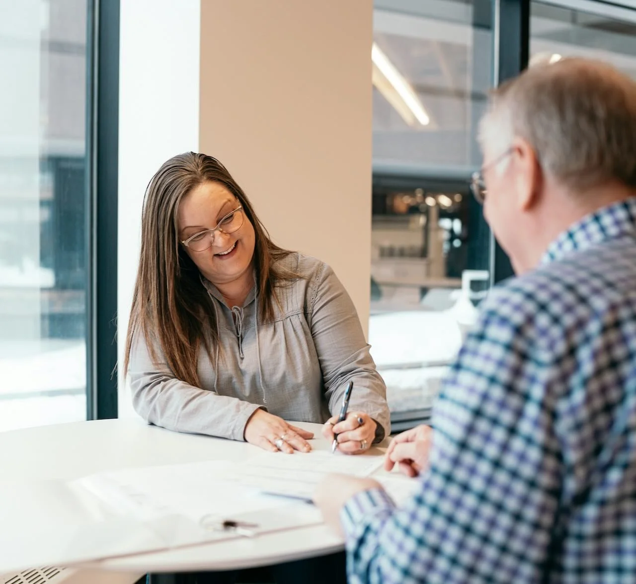 A woman and an elderly man sit at a white table in a bright office, engaging in a conversation while smiling.