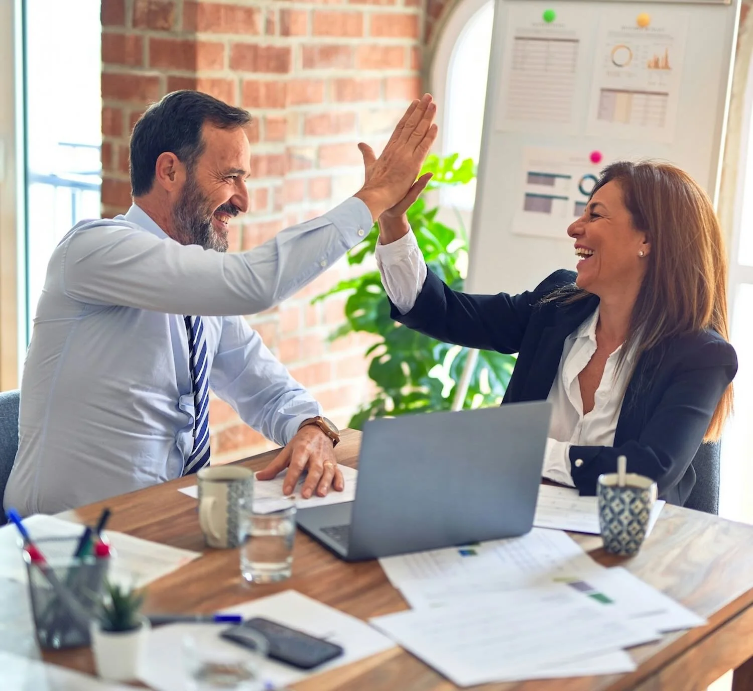 Two colleagues, a man and a woman, are smiling and high-fiving each other in a modern office. There are papers, a laptop, and office supplies on the desk, with a brick wall and a whiteboard in the background.