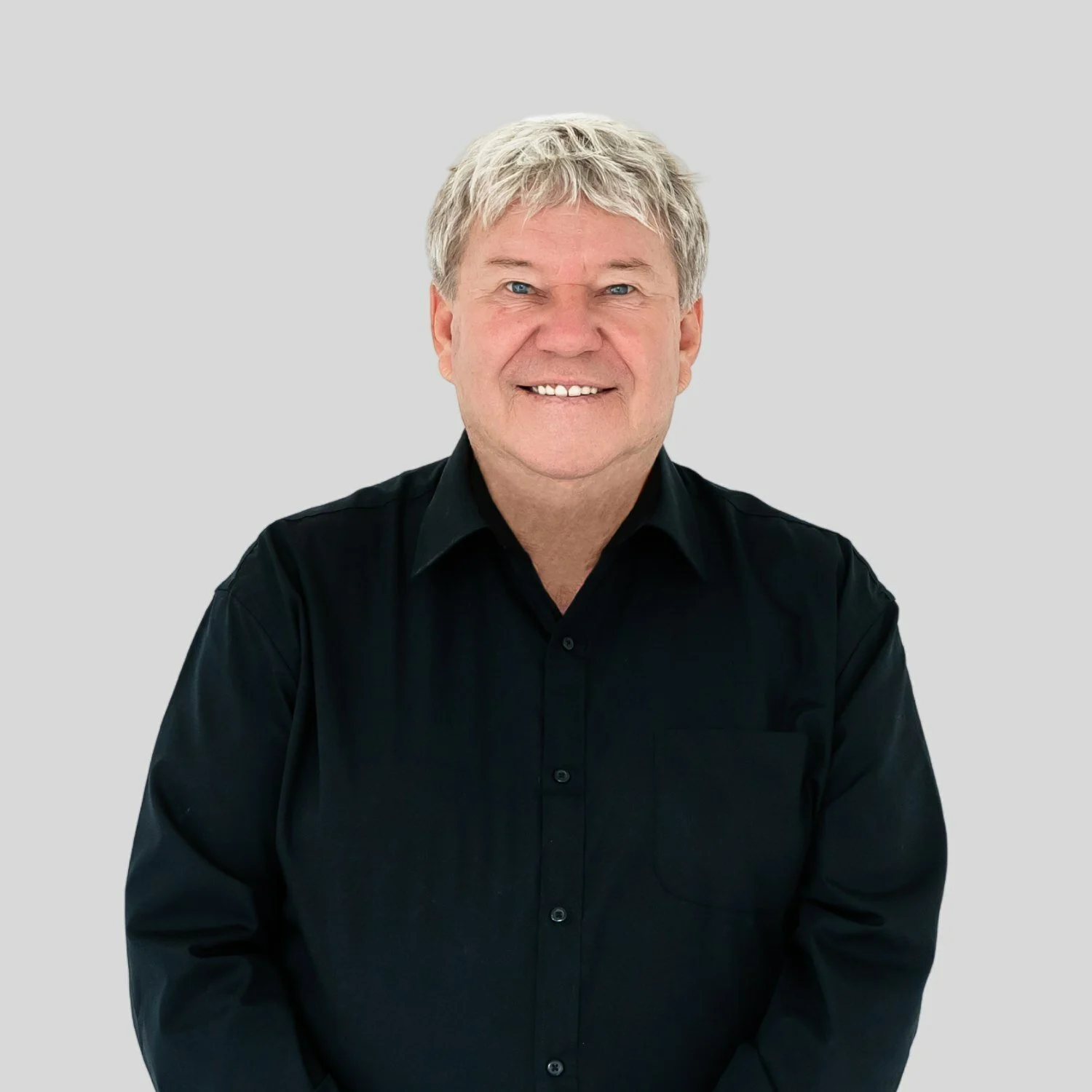A smiling man with gray hair wearing a black button-up shirt, standing against a plain light gray background.