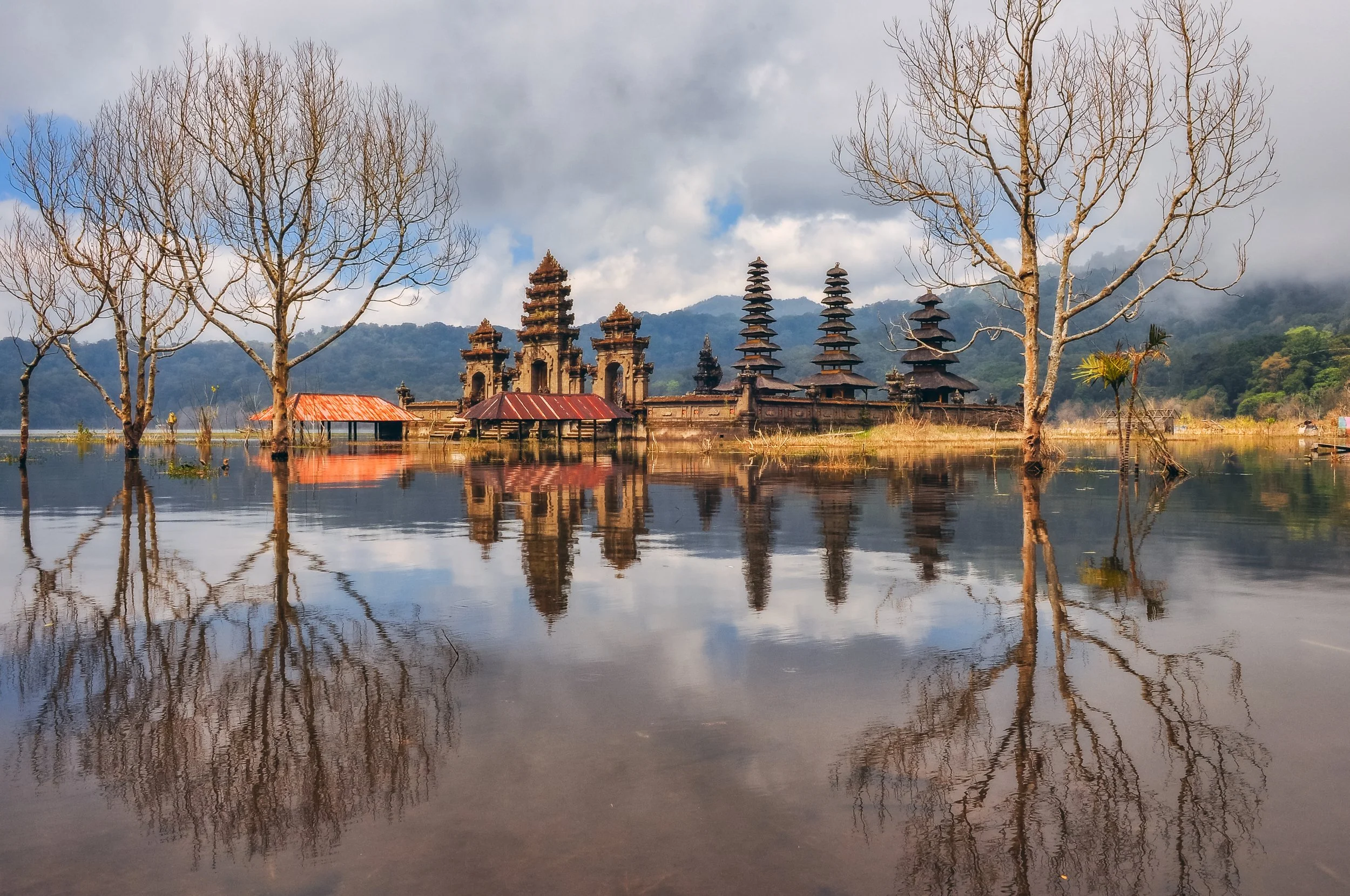 Paisaje con templos antiguos y árboles sin hojas frente a un lago, con montañas y nubes en el fondo.