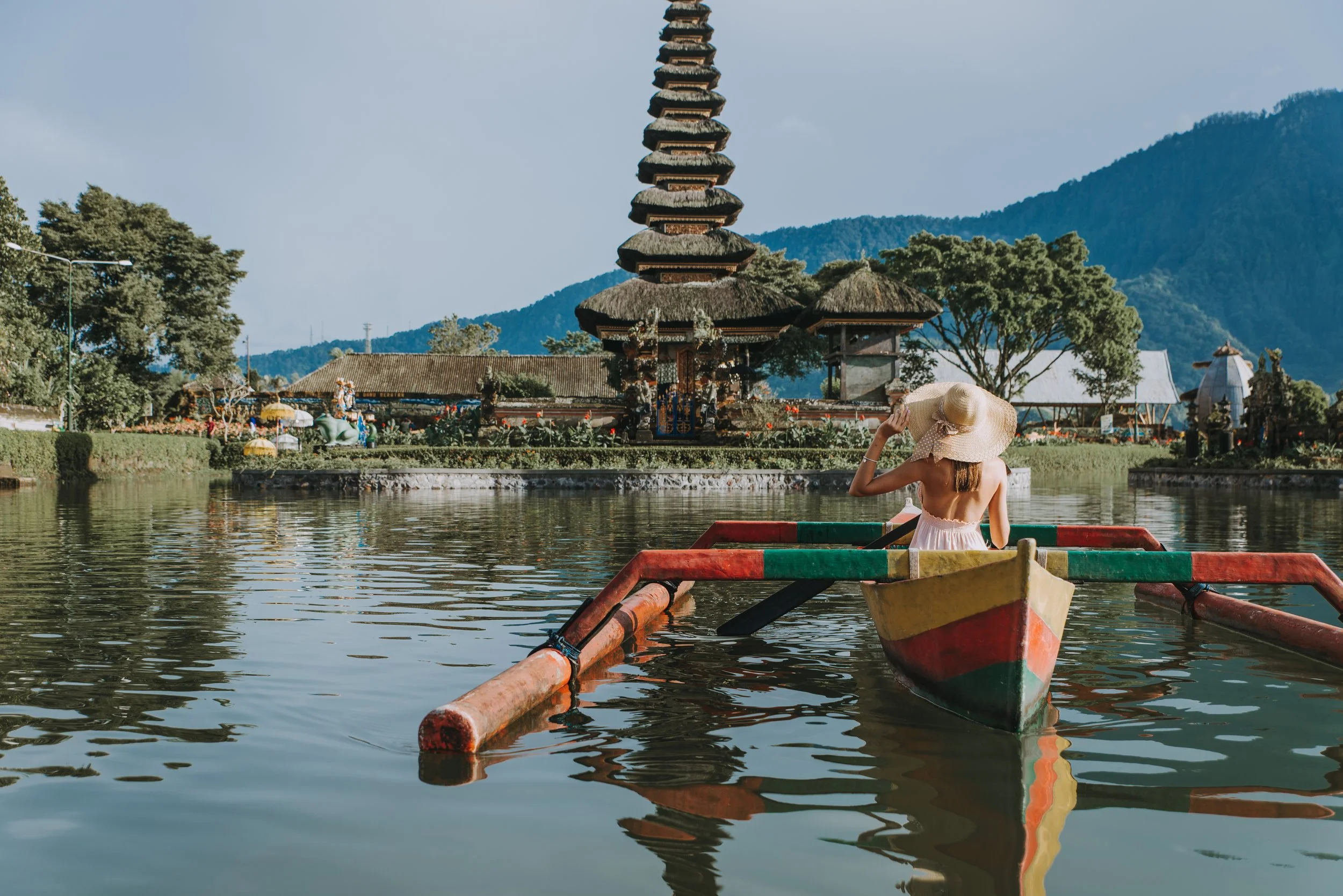 Una mujer con vestido rosado y sombrero grande en un bote de colores en un lago, con una pagoda en el fondo y montañas.