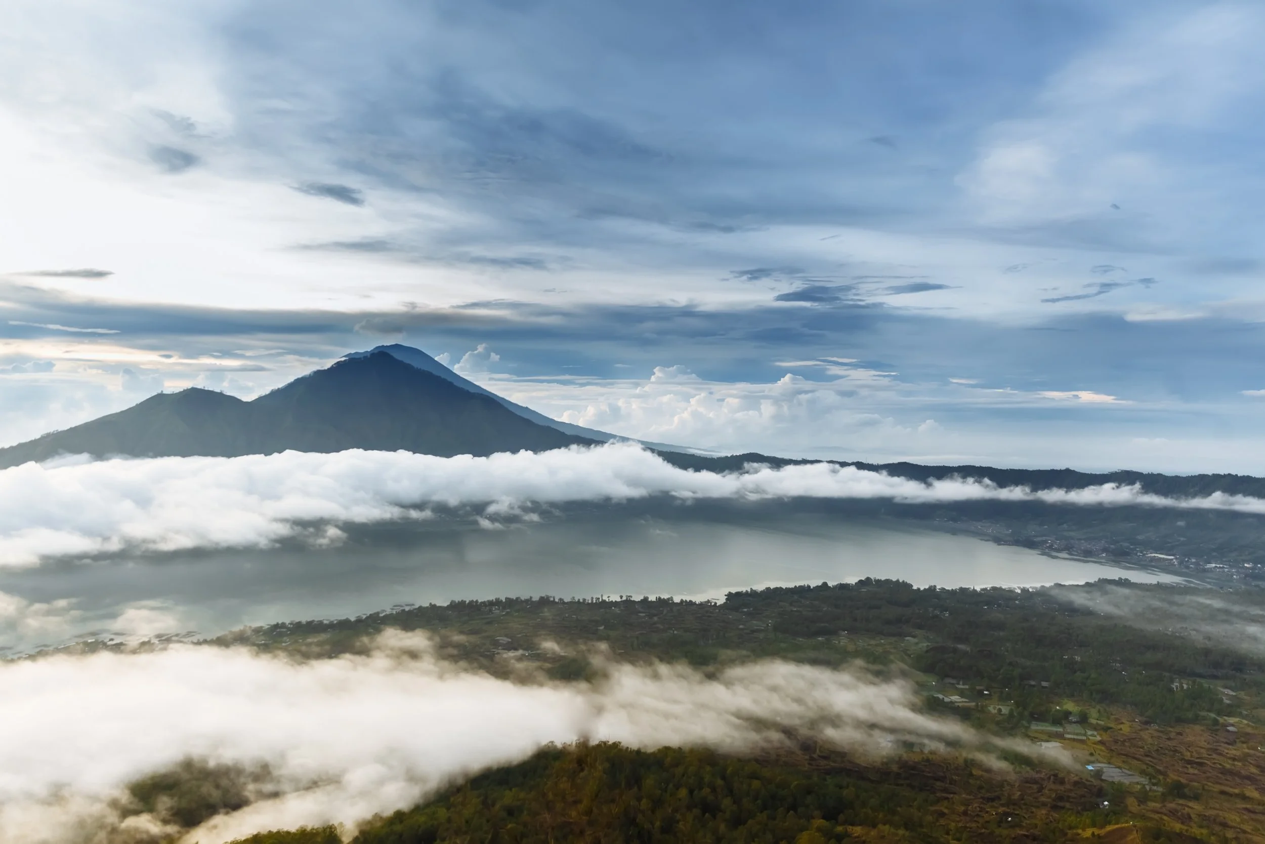 Vista panorámica de un volcán con nubosas en la base y en el cielo, y un lago junto a áreas de vegetación y agricultura.