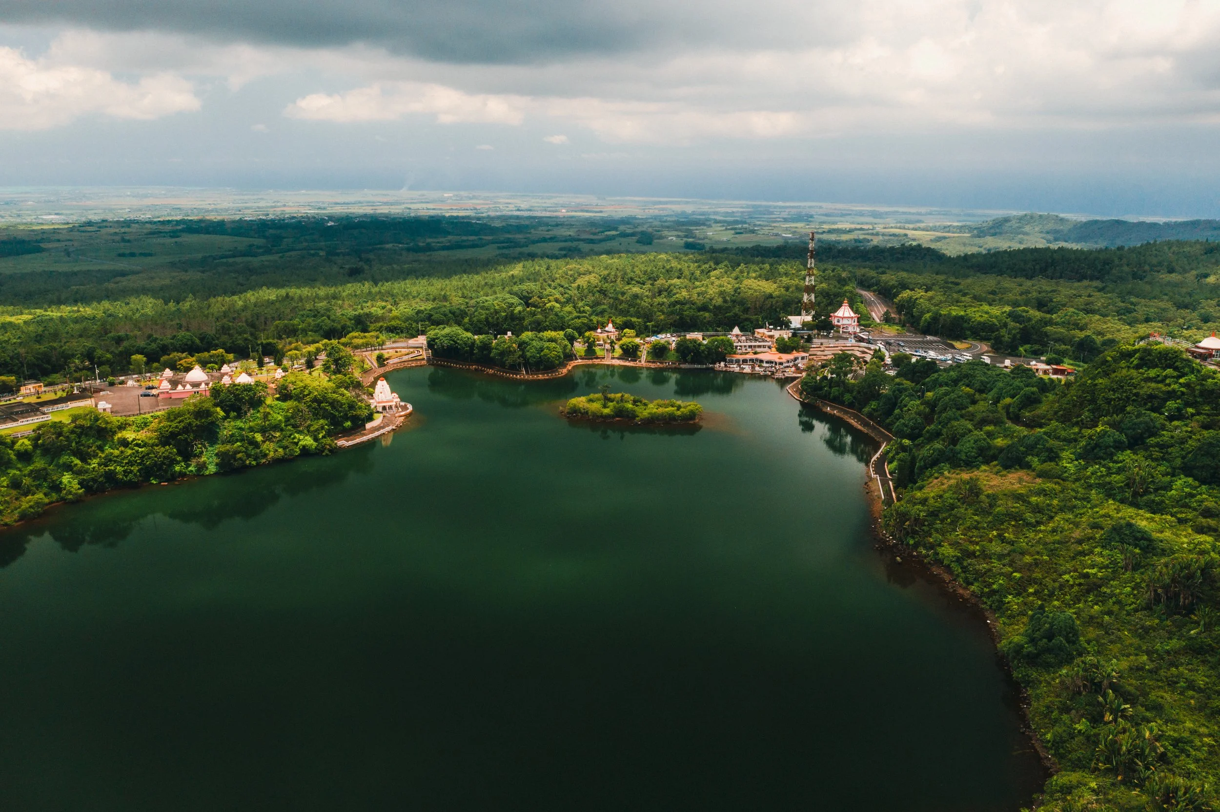 Vista aérea de un embalse rodeado de vegetación y árboles en un área montañosa, con edificios y una torre de telecomunicaciones en el fondo.