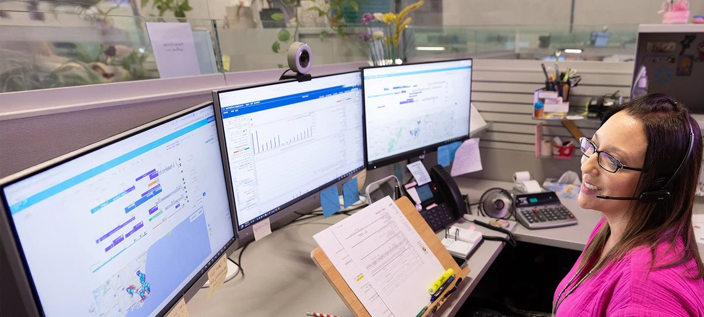 A woman wearing glasses and a pink shirt working at her cubicle in an office, with three computer monitors displaying charts, graphs, and maps, along with office supplies and personal items around her.