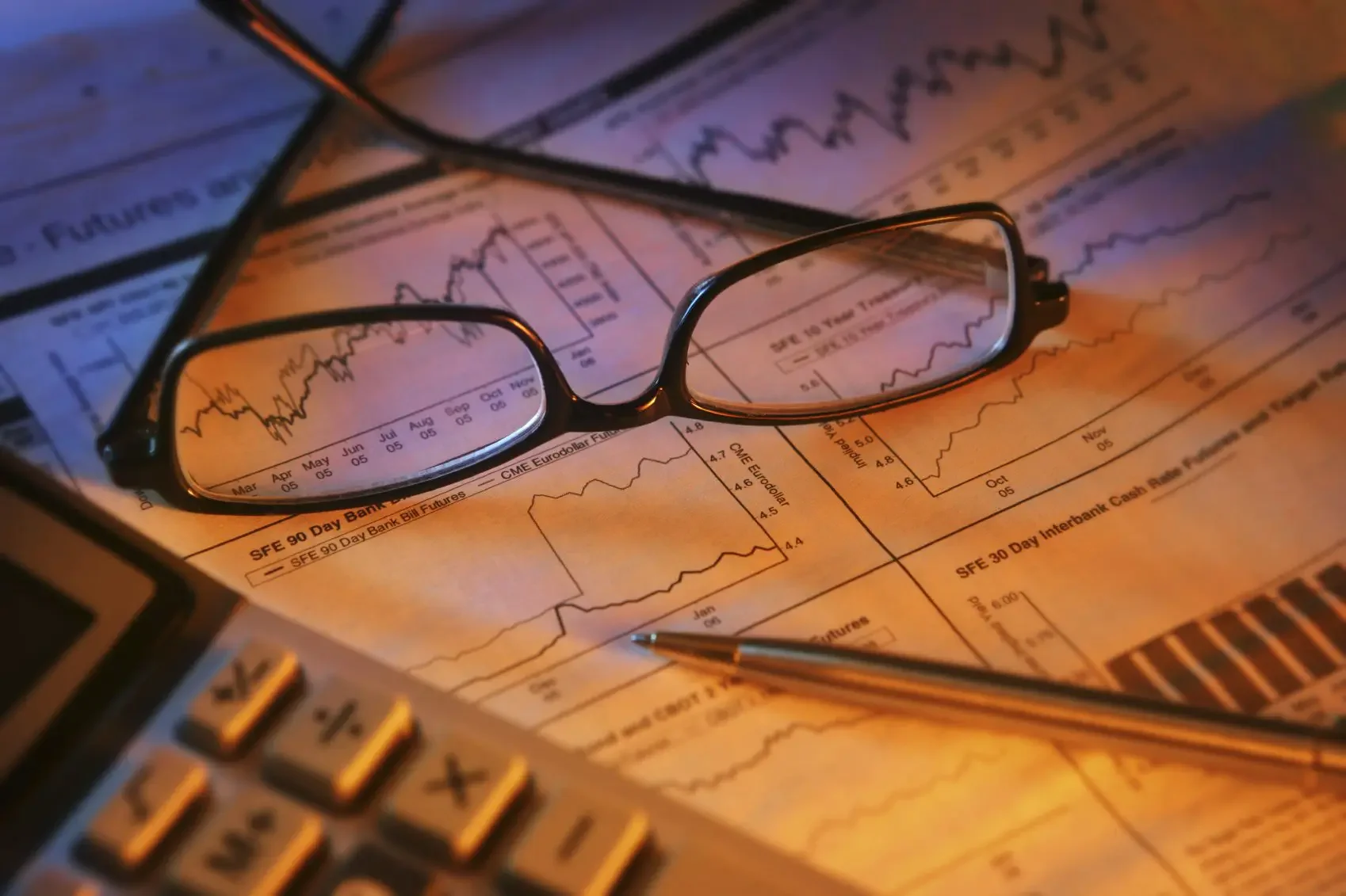 A pair of eyeglasses resting on financial charts and graphs on paper, with a pen and calculator nearby.