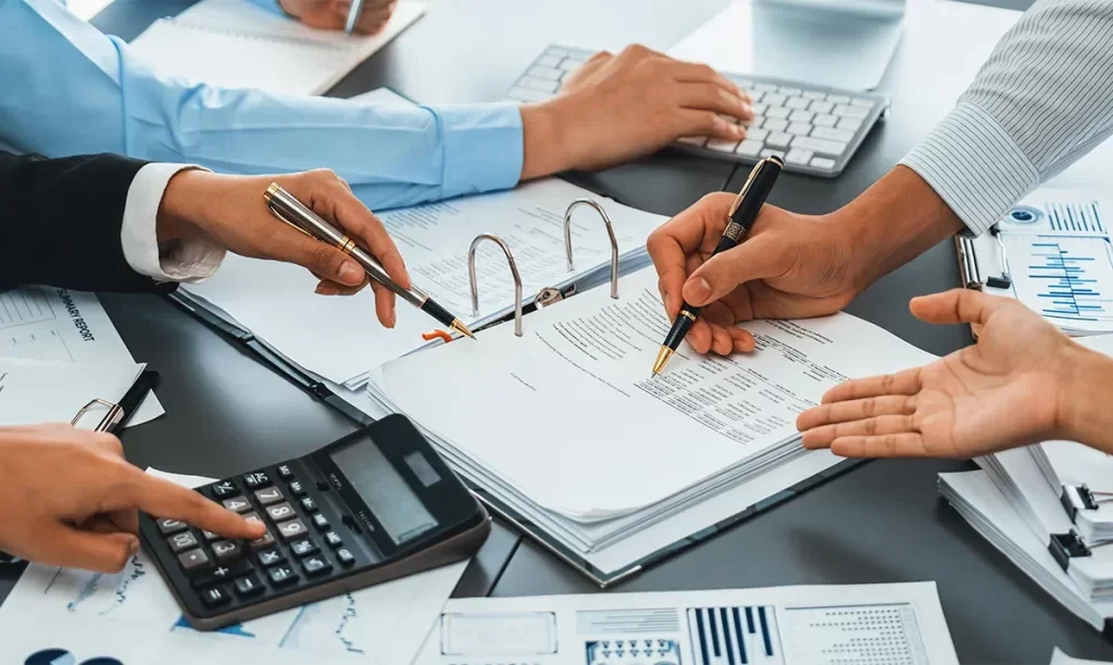 People working with financial documents, using pens and a calculator on a cluttered desk.