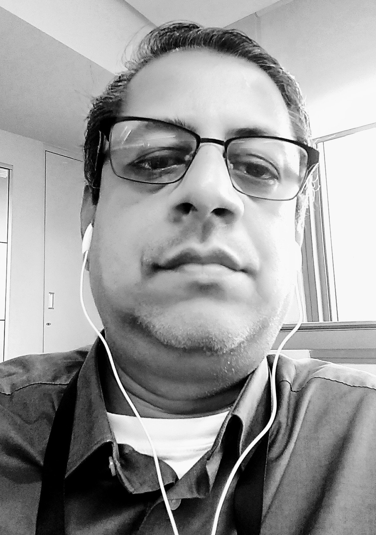 A black and white close-up photo of a man with glasses, earbuds, and a collared shirt, sitting indoors near a window.