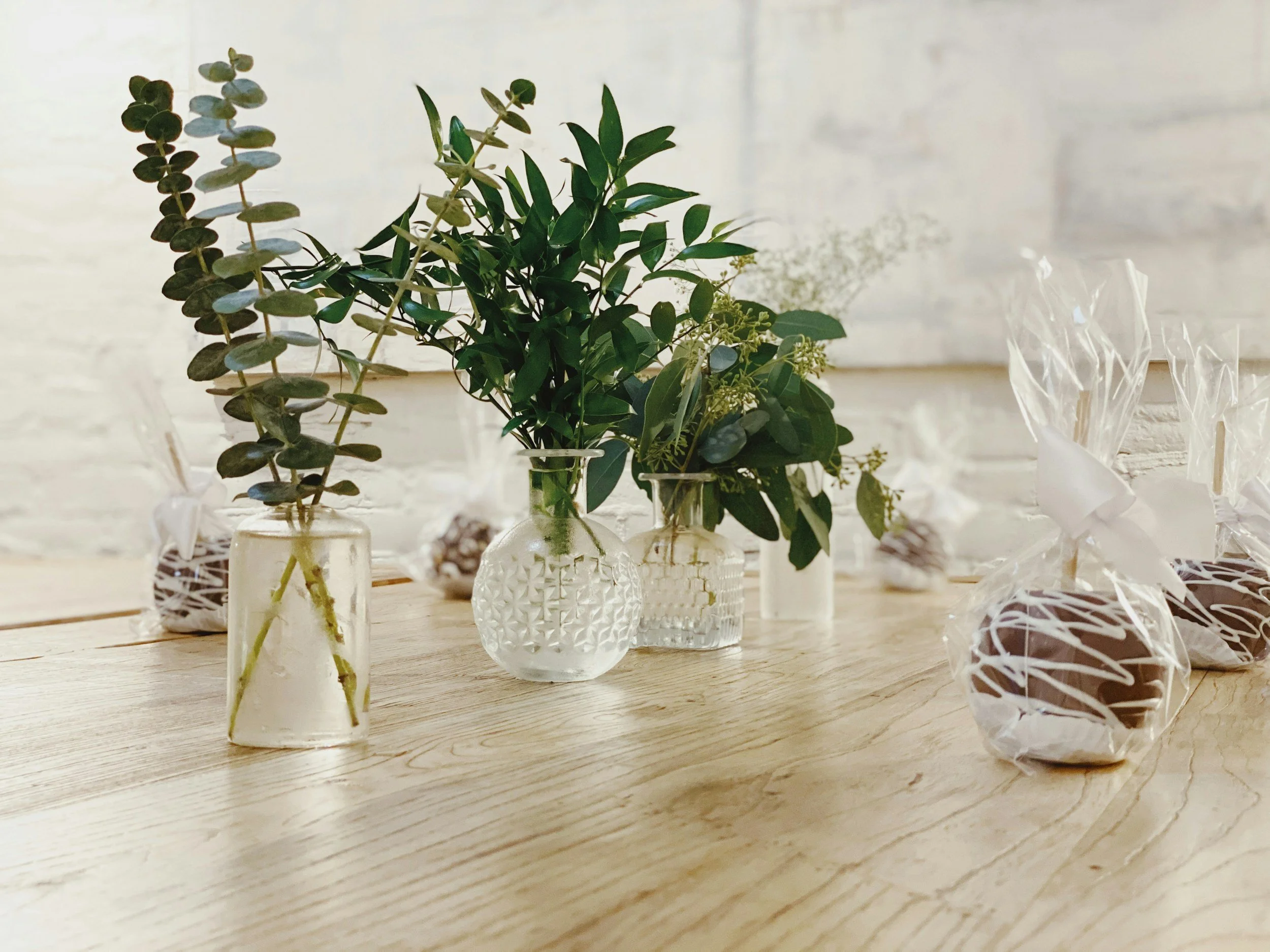 Three vases with green leaves and foliage on a wooden table, three wrapped chocolates in clear plastic with white ribbon at the side, in a room with white brick wall background.