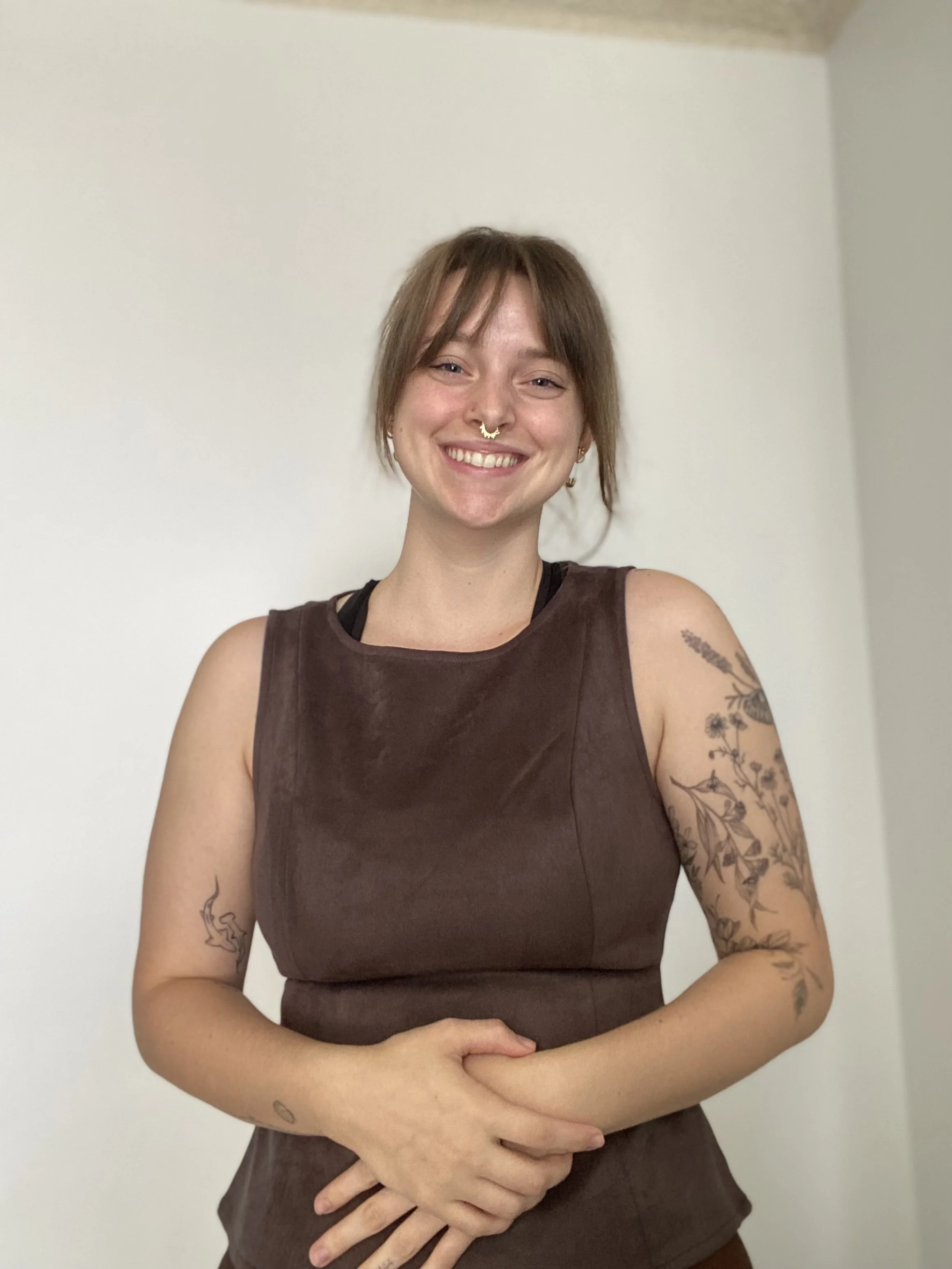 A smiling woman with short brown hair, tattoos on her arms, wearing a sleeveless brown top, standing against a plain beige wall.