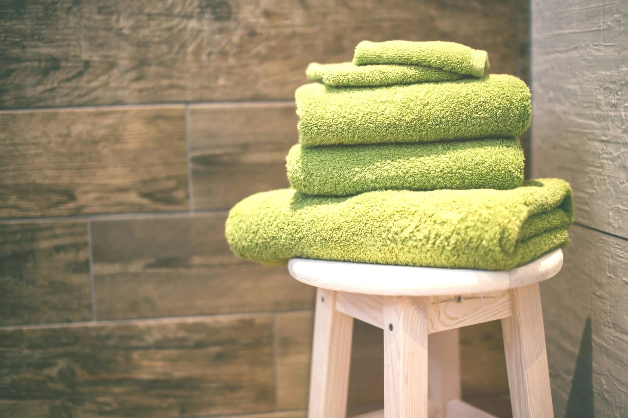 Four neatly folded green towels placed on a small wooden stool against a dark wooden wall background.