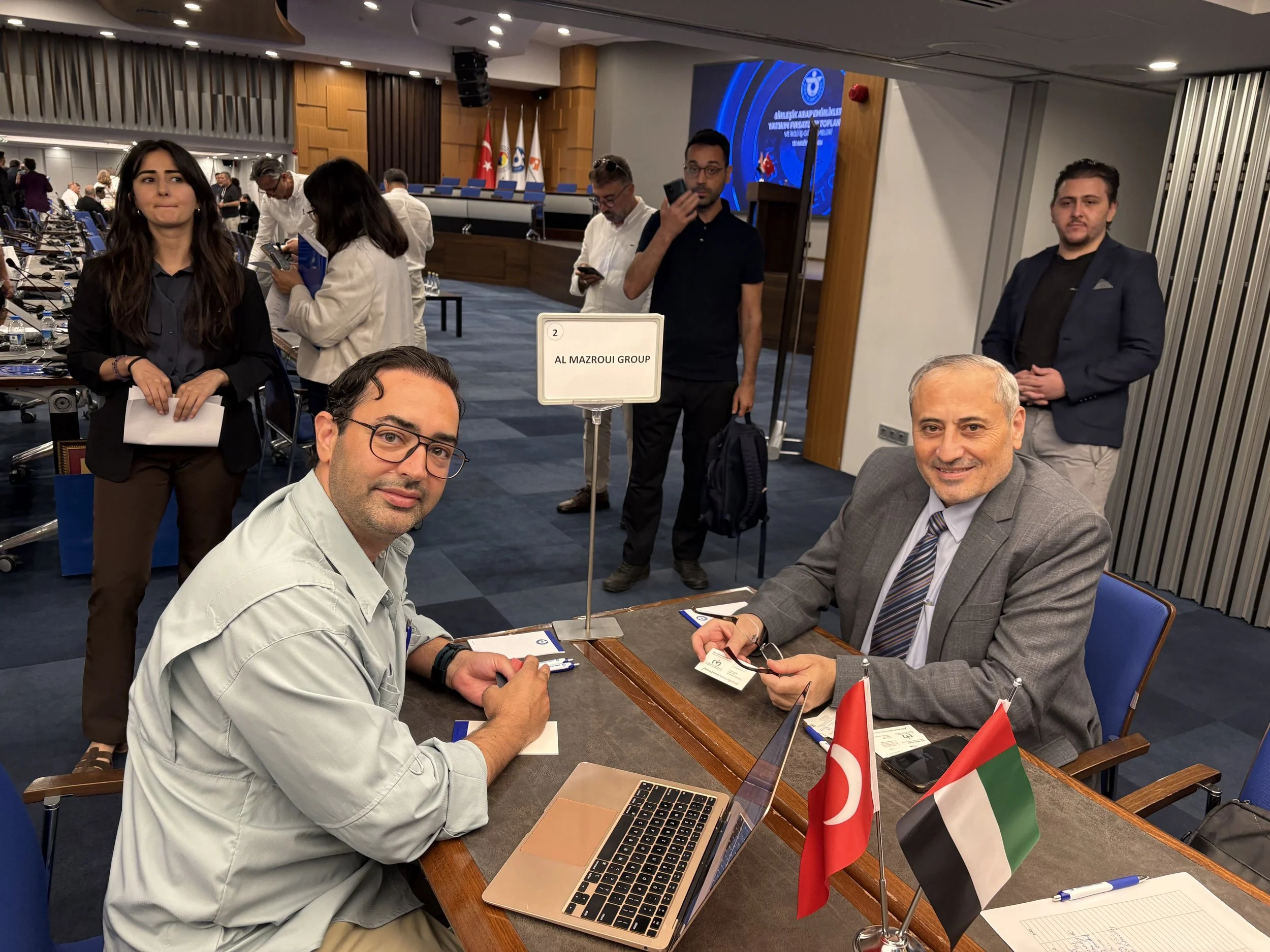 People attending a formal conference or meeting, some are seated at a table with flags of Turkey and Hungary, while others stand or sit in the background. The room is decorated with flags, banners, and a large screen.