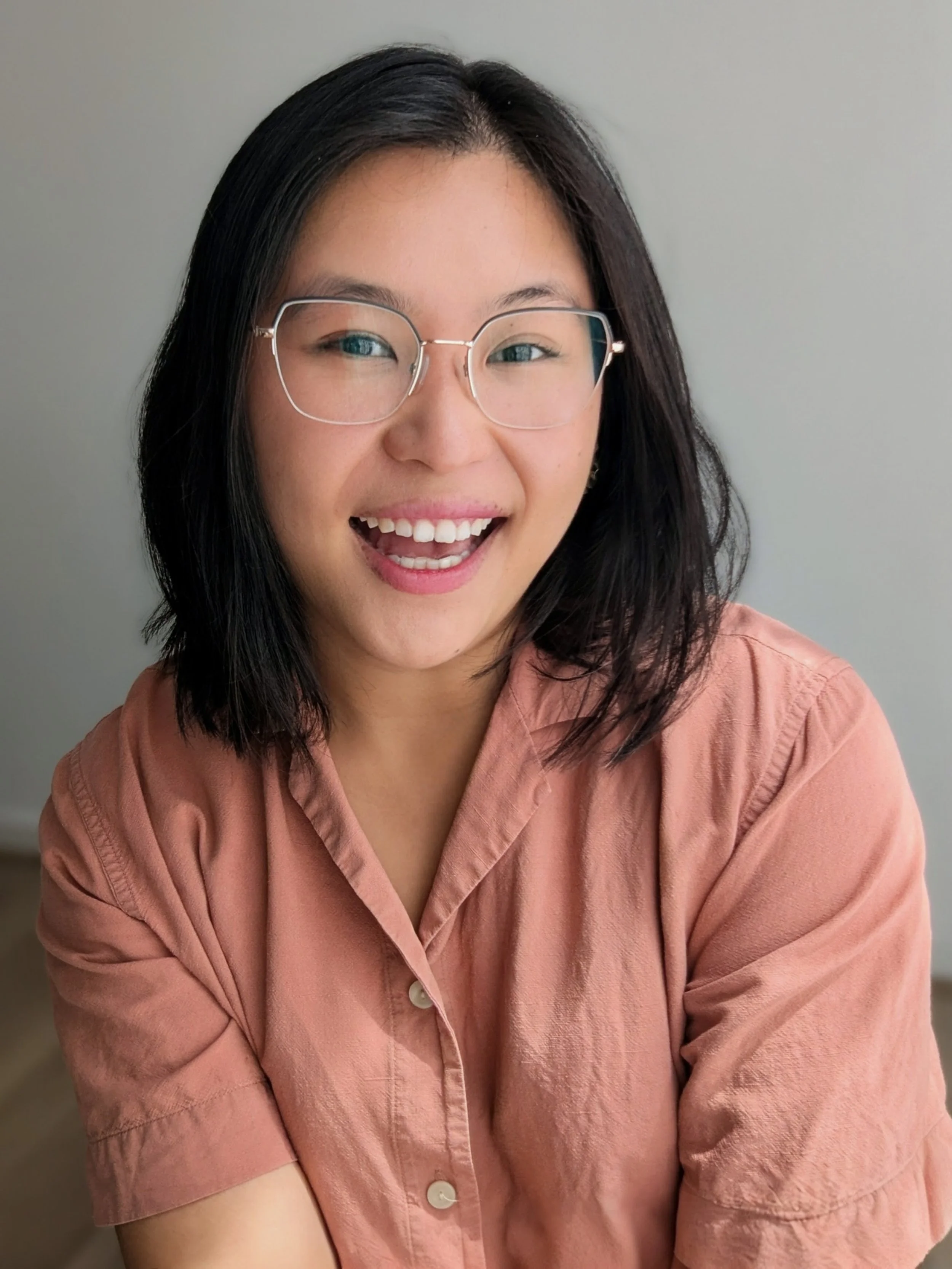 A young woman with dark shoulder-length hair, wearing glasses and a pinkish-brown button-up shirt, smiling.