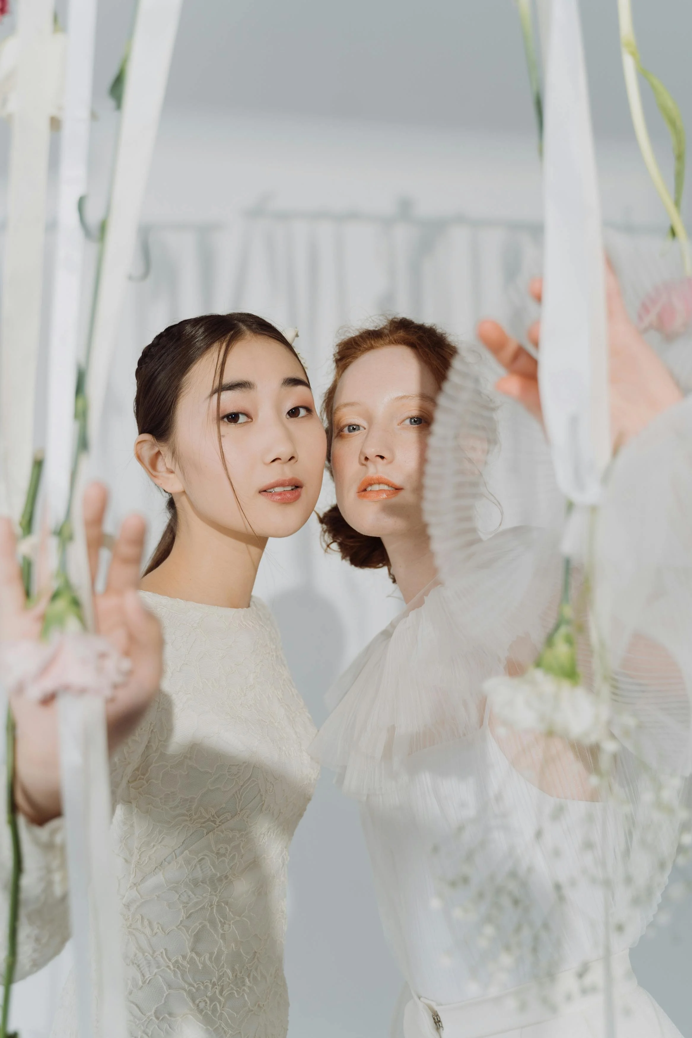 Two women with different ethnic backgrounds take a selfie together, framed by a white floral structure, against a soft, light-colored background.