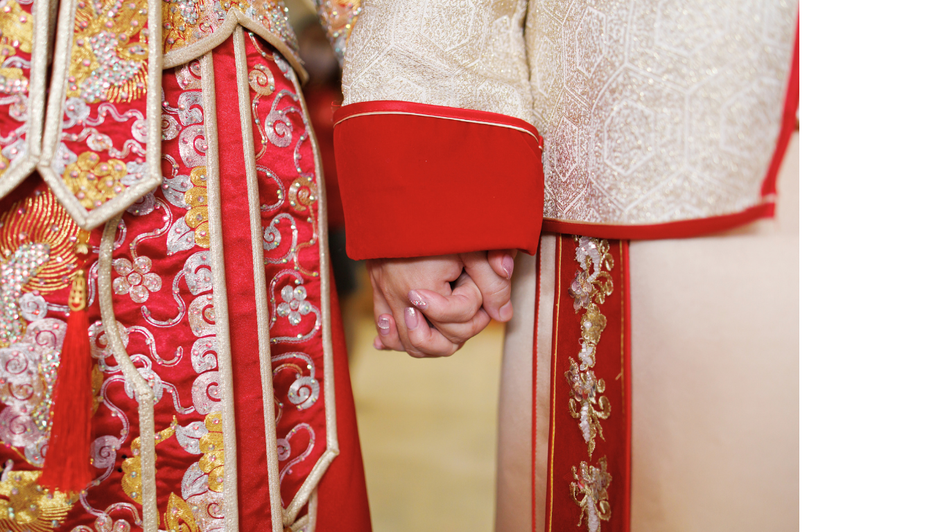 Close-up of two people holding hands, wearing traditional Asian wedding attire with intricate embroidery, red and gold colors, and detailed patterns.