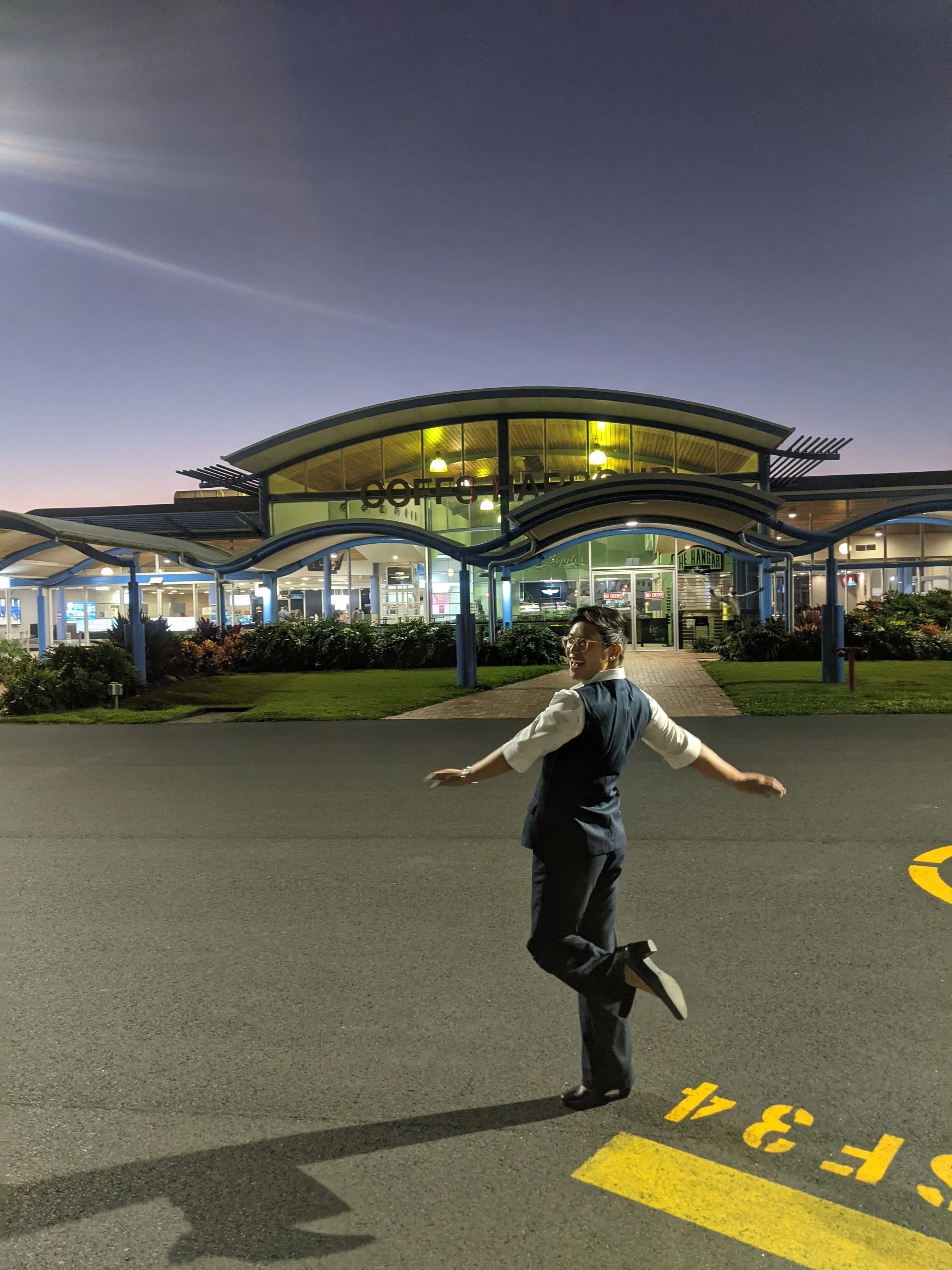 A person in a suit striking a playful pose on a parking lot at twilight, with a modern building in the background.