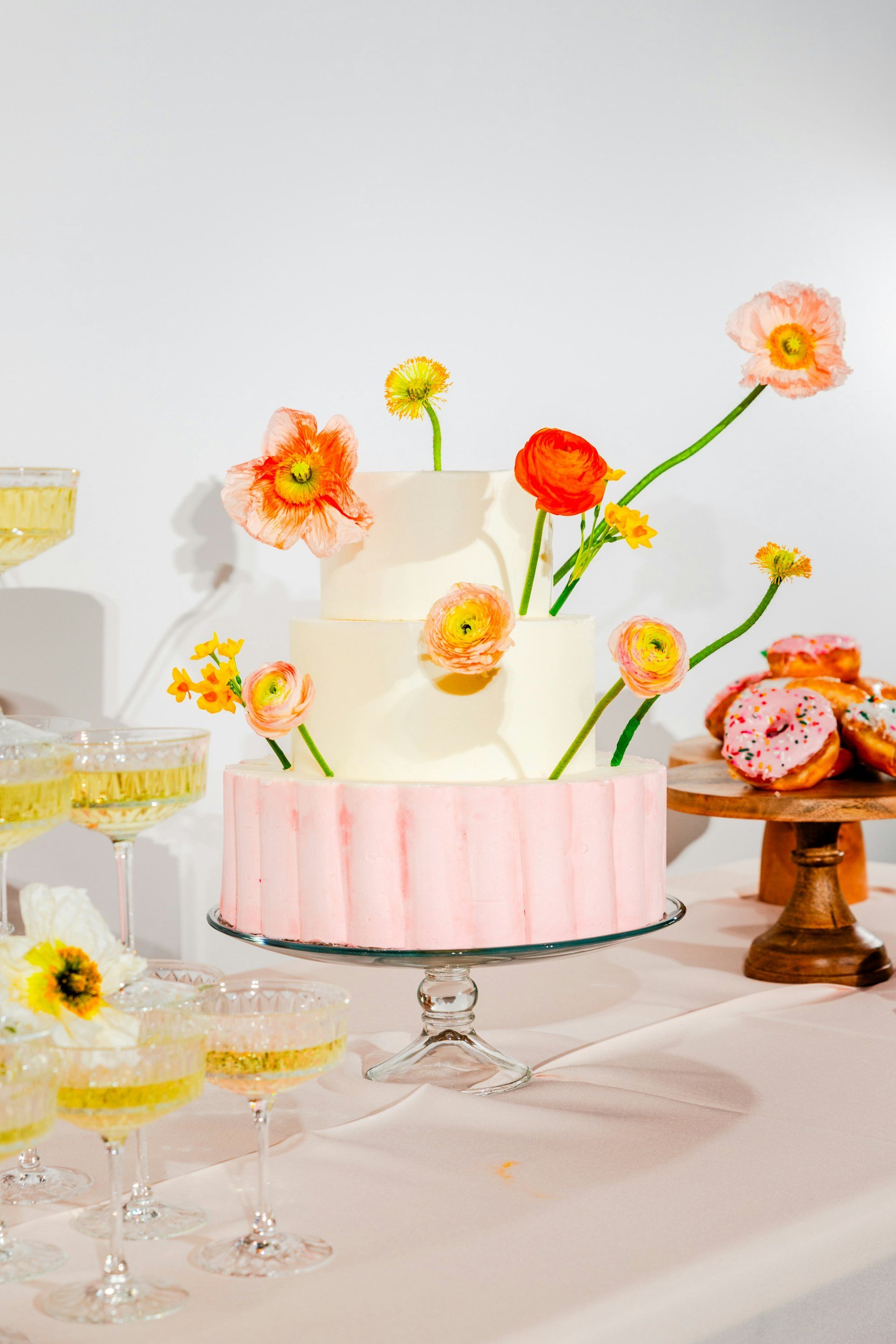 A colorful, three-tiered birthday cake decorated with edible flowers and positioned on a glass cake stand, surrounded by champagne glasses, and pink donuts with sprinkles on a wooden tray, all on a light pink tablecloth.