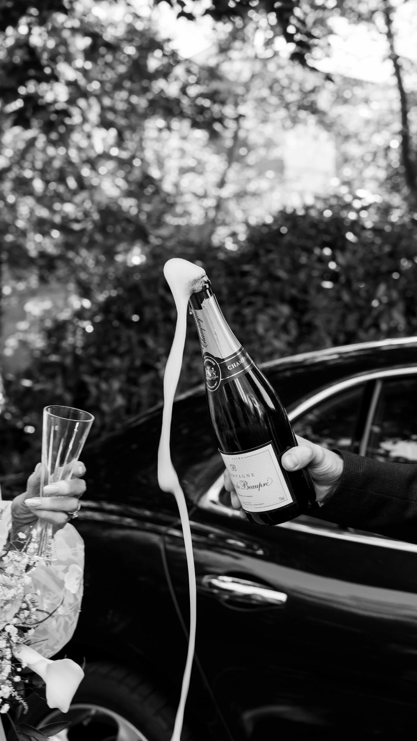 Two people celebrating, one holding a champagne bottle with foam pouring out, and the other holding a champagne glass, near a black car with a background of trees.
