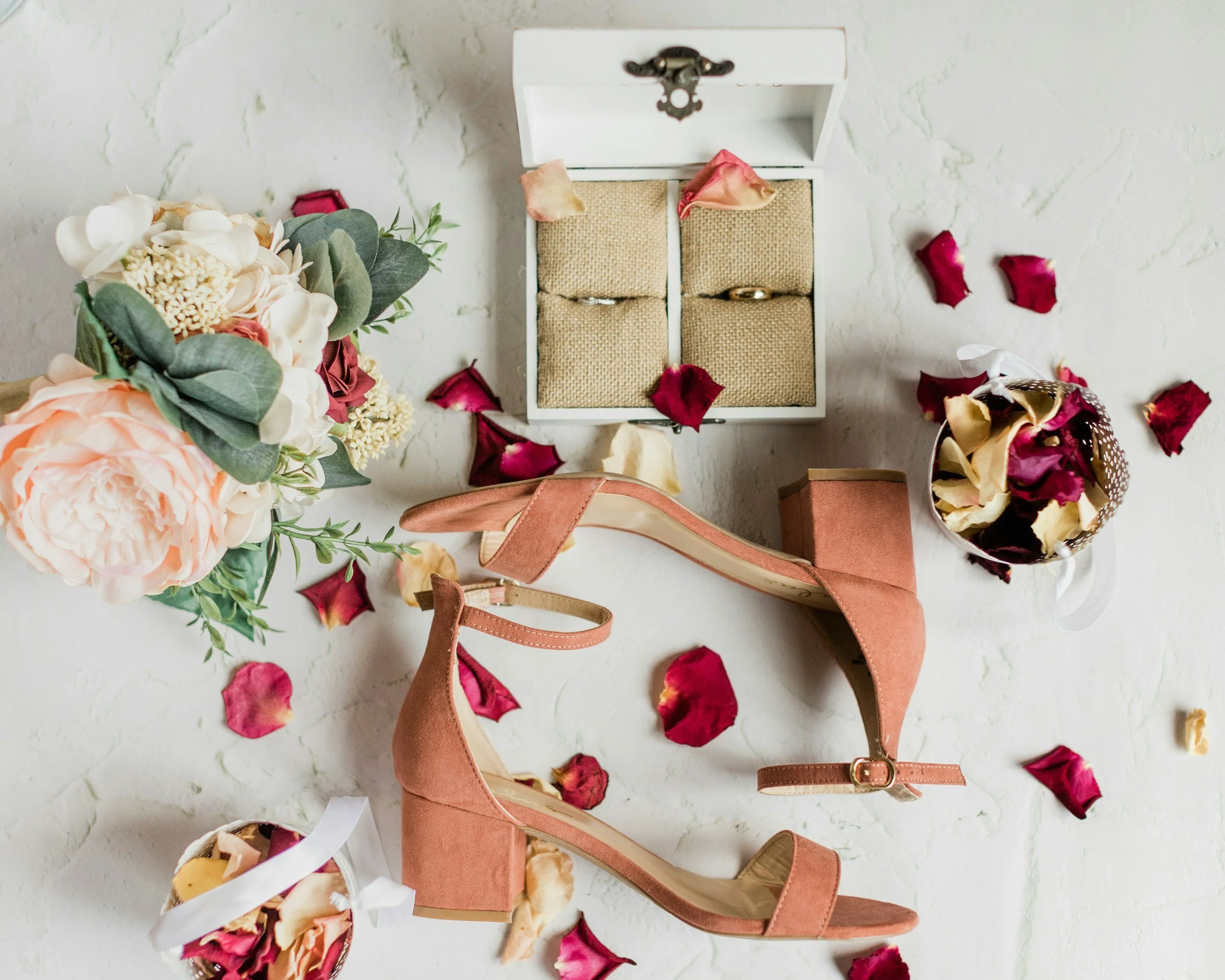 A pair of pink high-heeled shoes surrounded by flower arrangements, a box of rings, and scattered rose petals on a white surface.