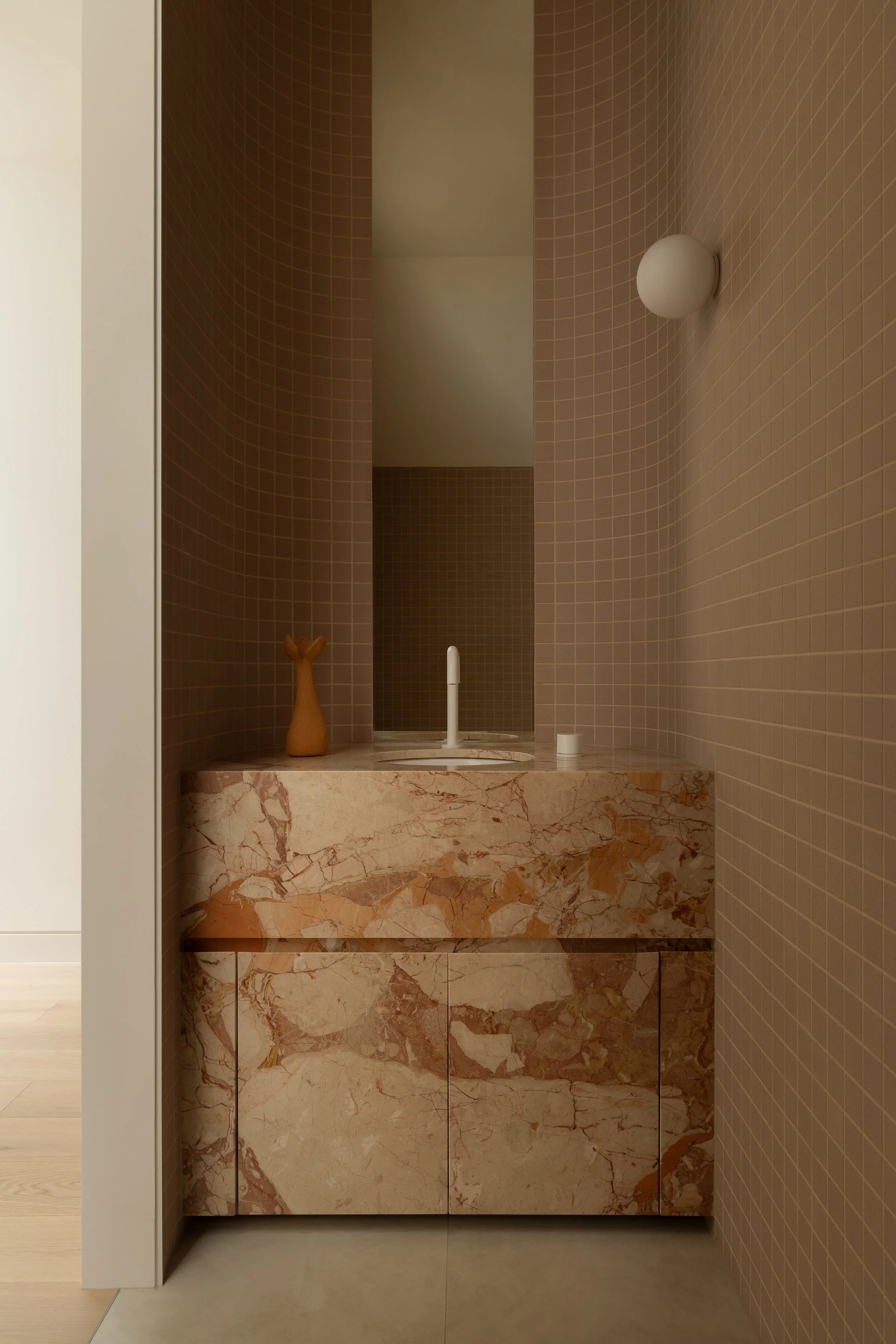A bathroom vanity with a marble surface, beige doors, a small wooden deer sculpture, a white faucet, a wall-mounted white light fixture, beige tiled walls, and a mirror reflecting the room.