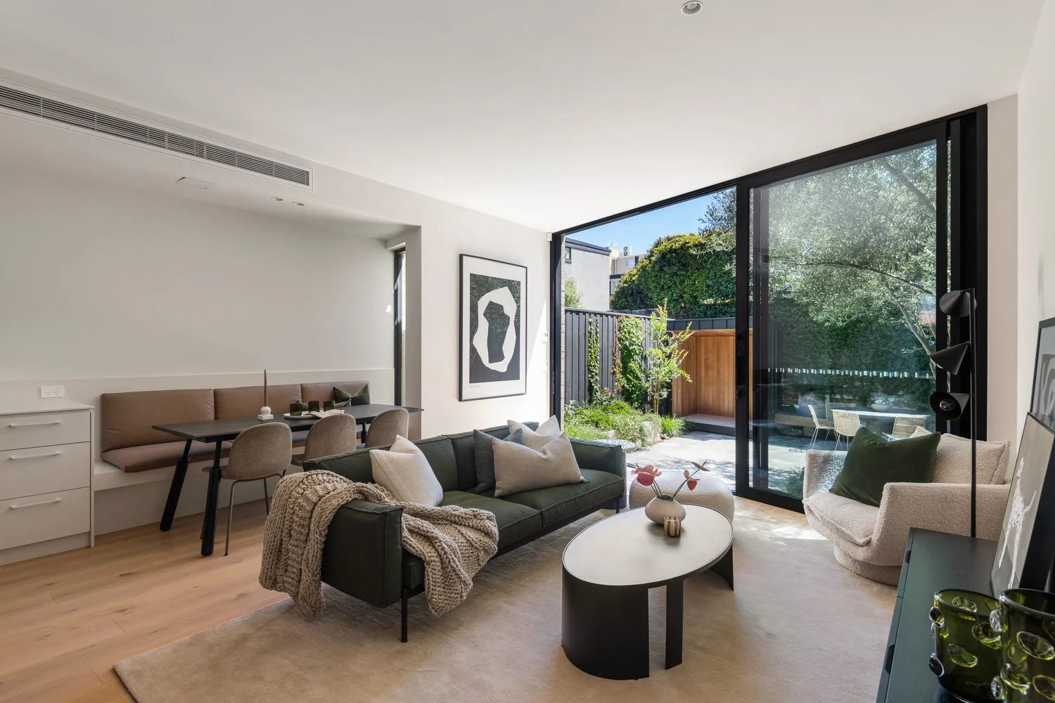 Modern living room with a black sofa, armchair, and oval coffee table, opening to a lush outdoor garden through large glass doors.