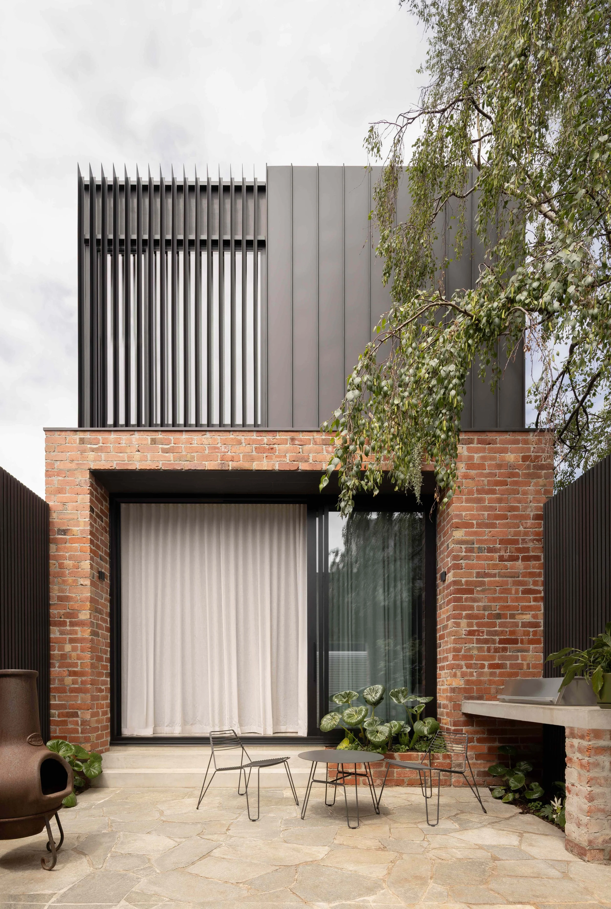 Modern house with brick and metal exterior, outdoor patio with furniture, and greenery.