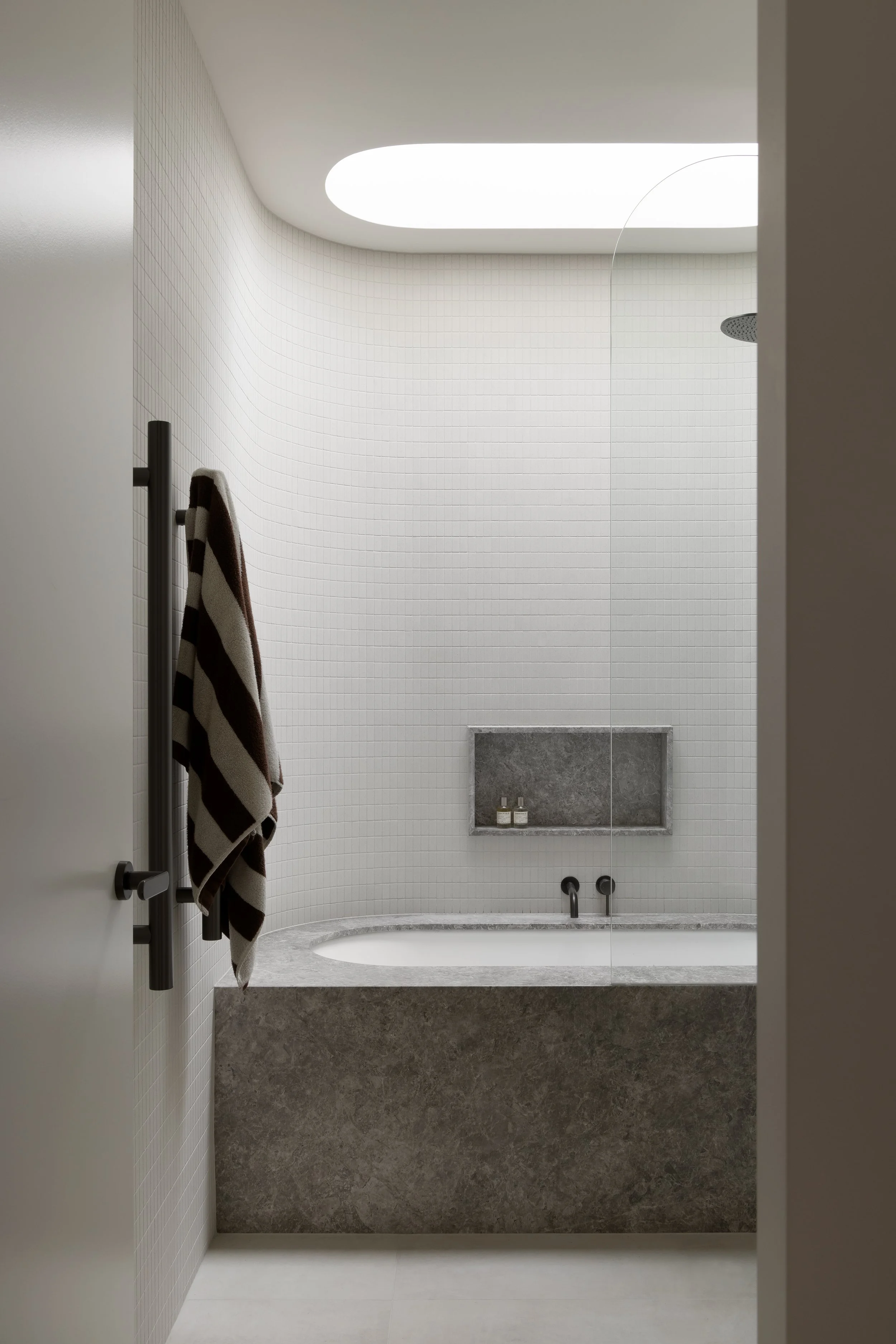Minimalist bathroom with a built-in bathtub, wood-textured wall niche, round skylight ceiling, and a towel hanging on a black towel rack.