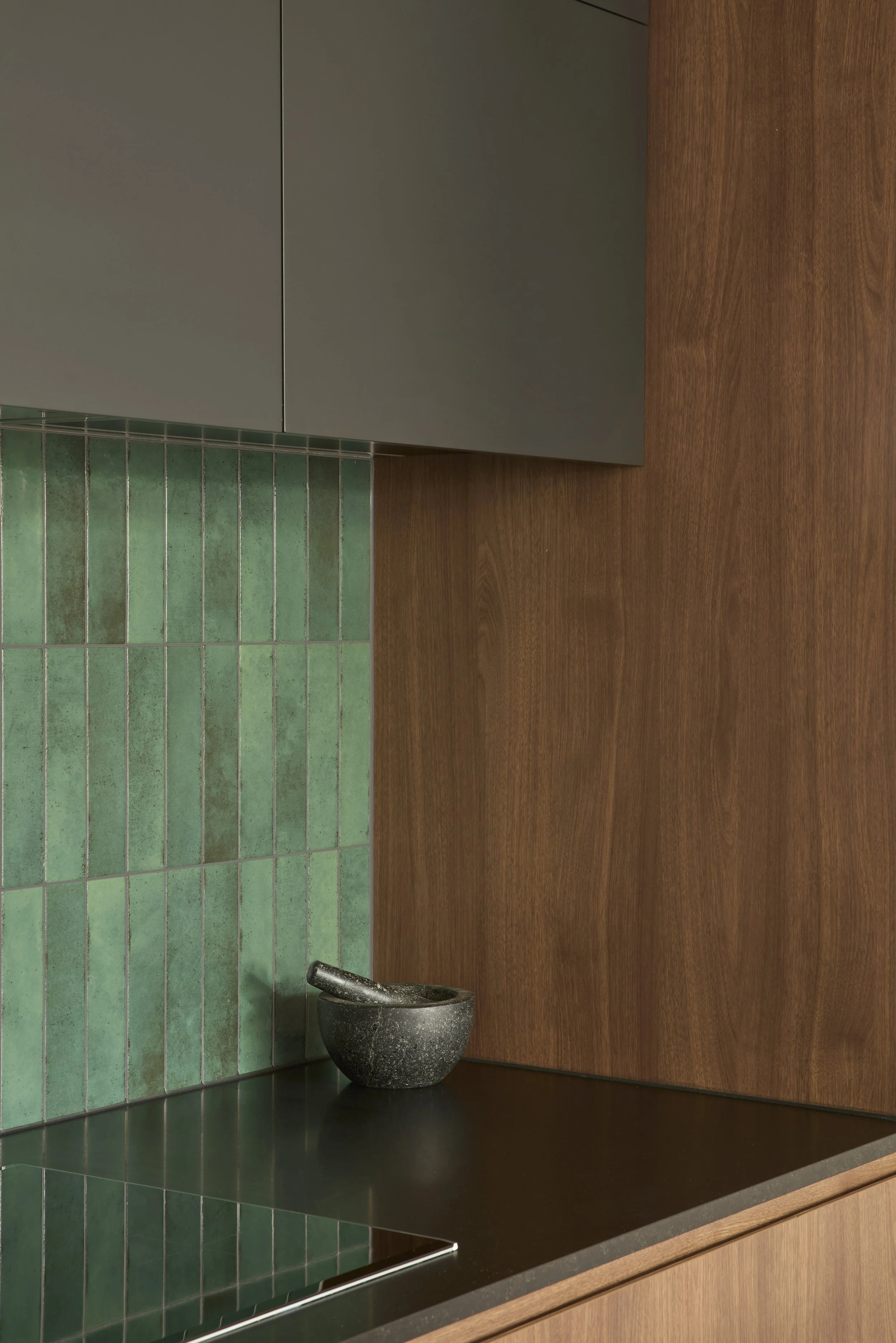 Close-up of a kitchen countertop with green tiles, wooden cabinetry, and a black mortar and pestle.