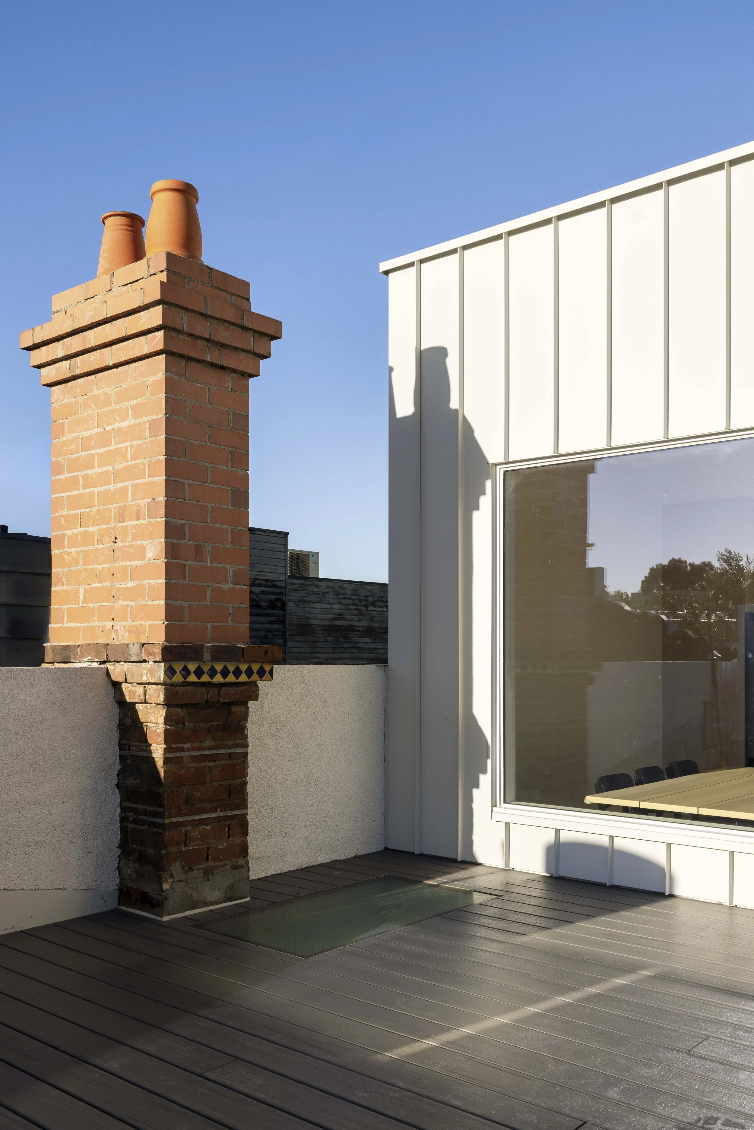 A rooftop terrace featuring a brick chimney, adjacent to a modern white building with large window, under a clear blue sky.