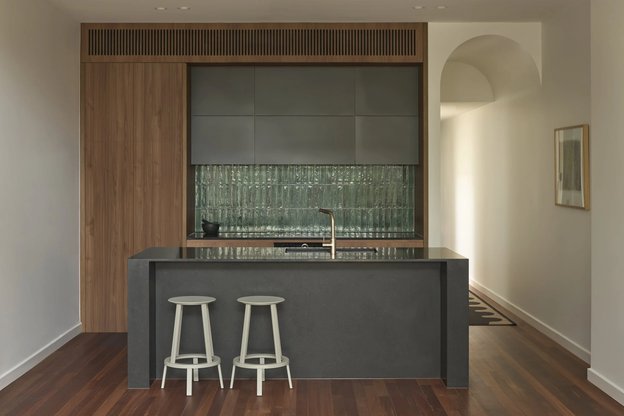 Modern kitchen with black island, two white stools, gray cabinets, green tiled backsplash, and wood accents.
