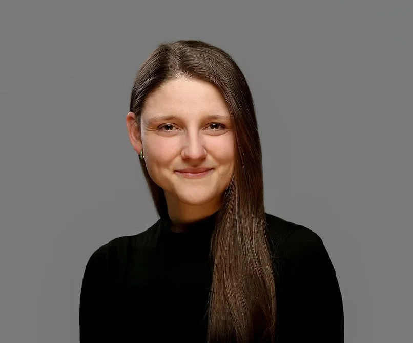 A woman with long brown hair wearing a black top, smiling and looking at the camera against a gray background.