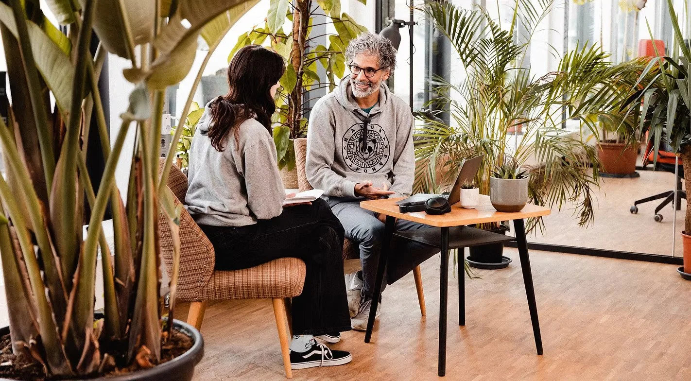 Two people sitting in a cozy indoor garden or plant-filled office, engaging in a conversation with smiles. One person appears to be showing notes, while the other listens. A laptop and potted plants are on the table.