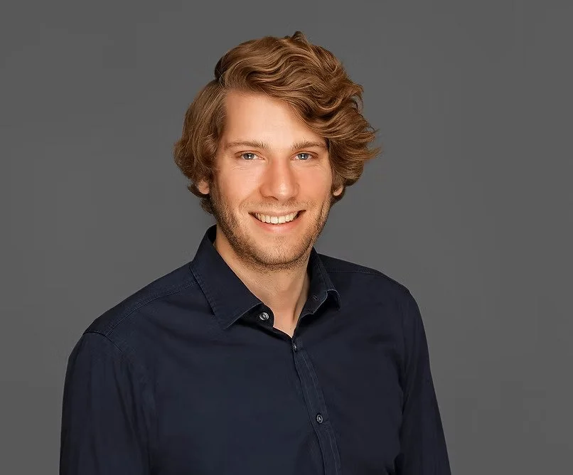 A smiling man with wavy blond hair wearing a dark blue collared shirt against a gray background.