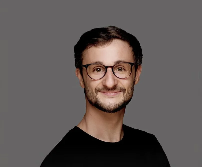 Headshot of a man with glasses, short dark hair, and a beard, smiling against a gray background.