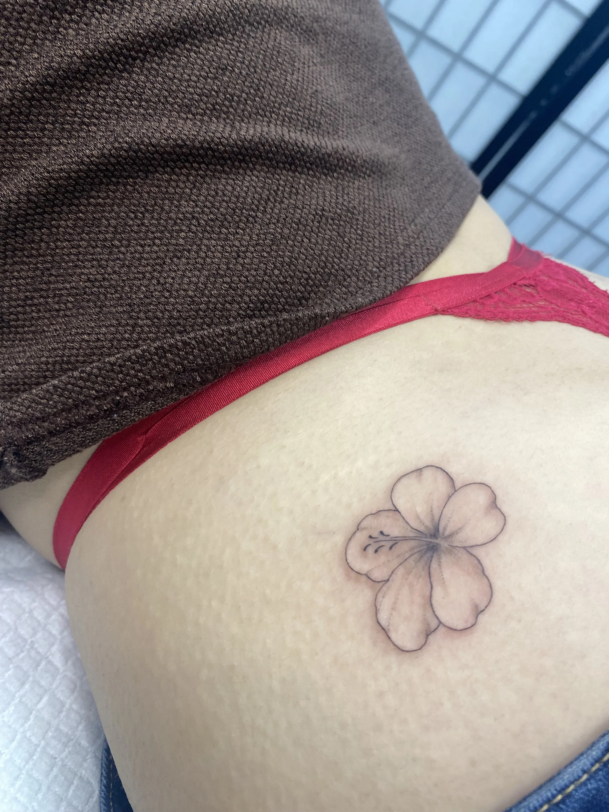 Close-up of a person's hip with a floral tattoo, wearing red lace underwear and a brown top, with a background of a tiled wall.