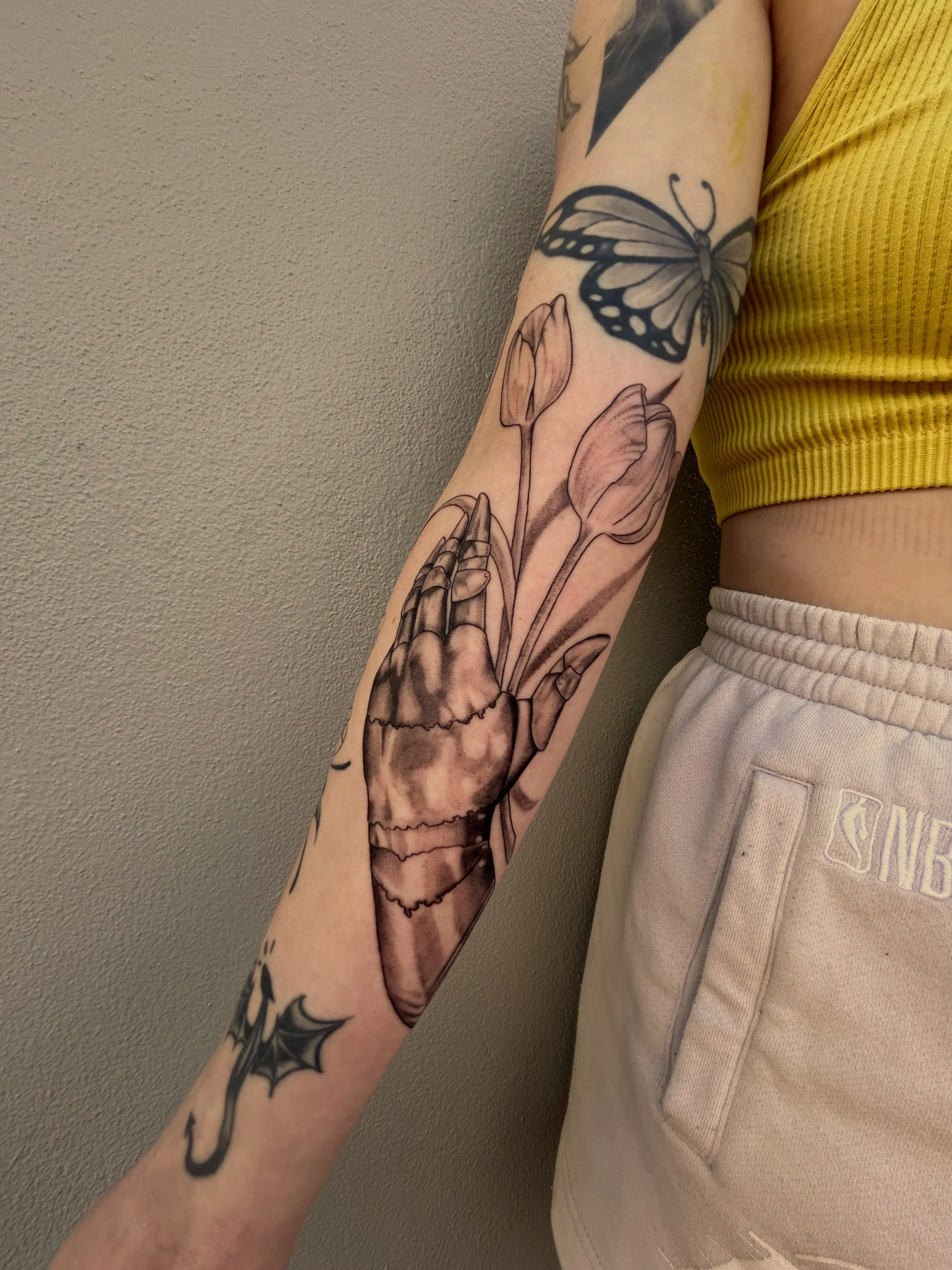 Tattooed arm with a butterfly, tulips, praying hands, and a bat with a moon on the wrist, wearing a yellow crop top and beige shorts.