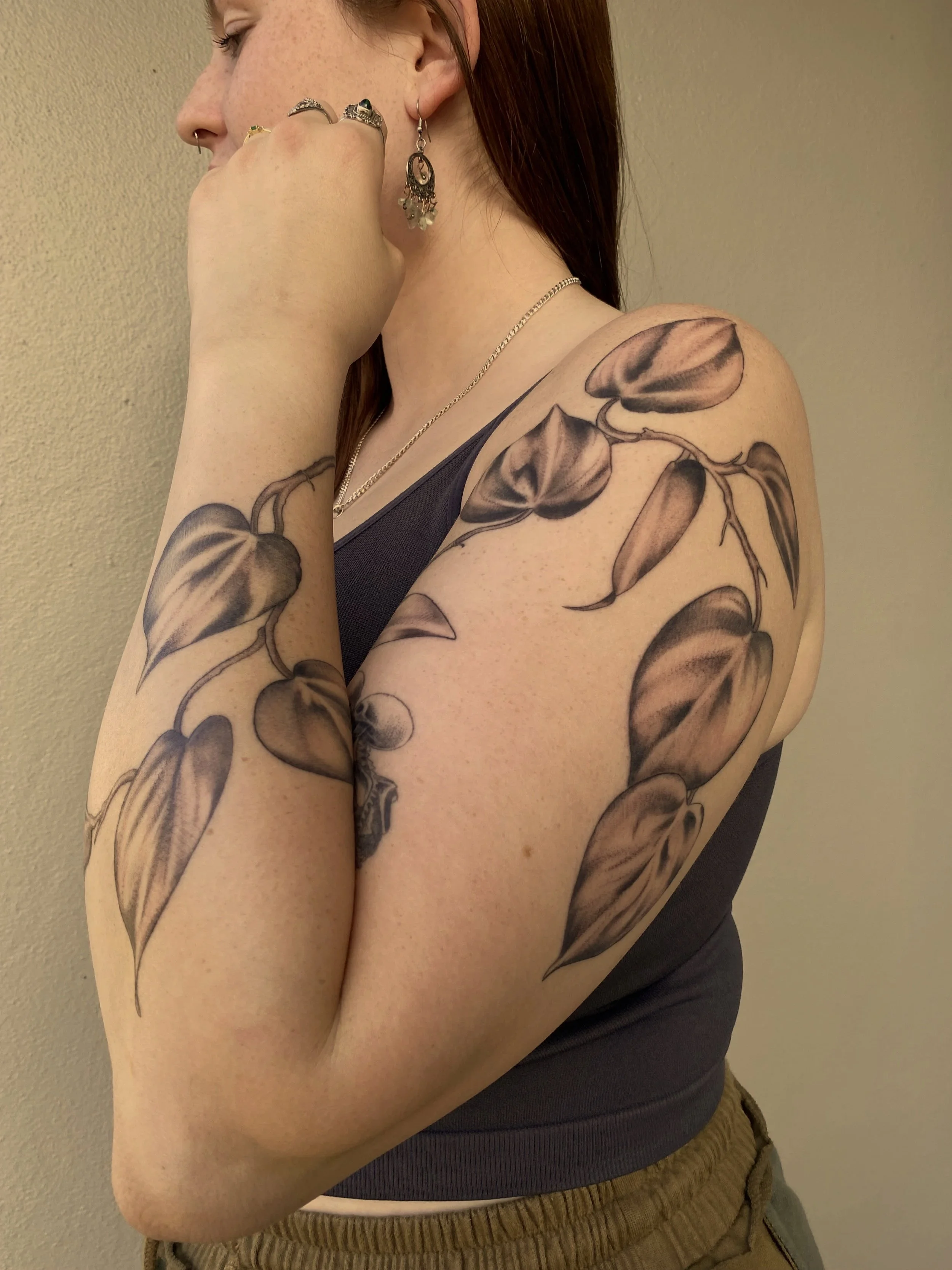 Close-up of a woman with a floral tattoo on her arm, wearing jewelry and a dark sleeveless top.