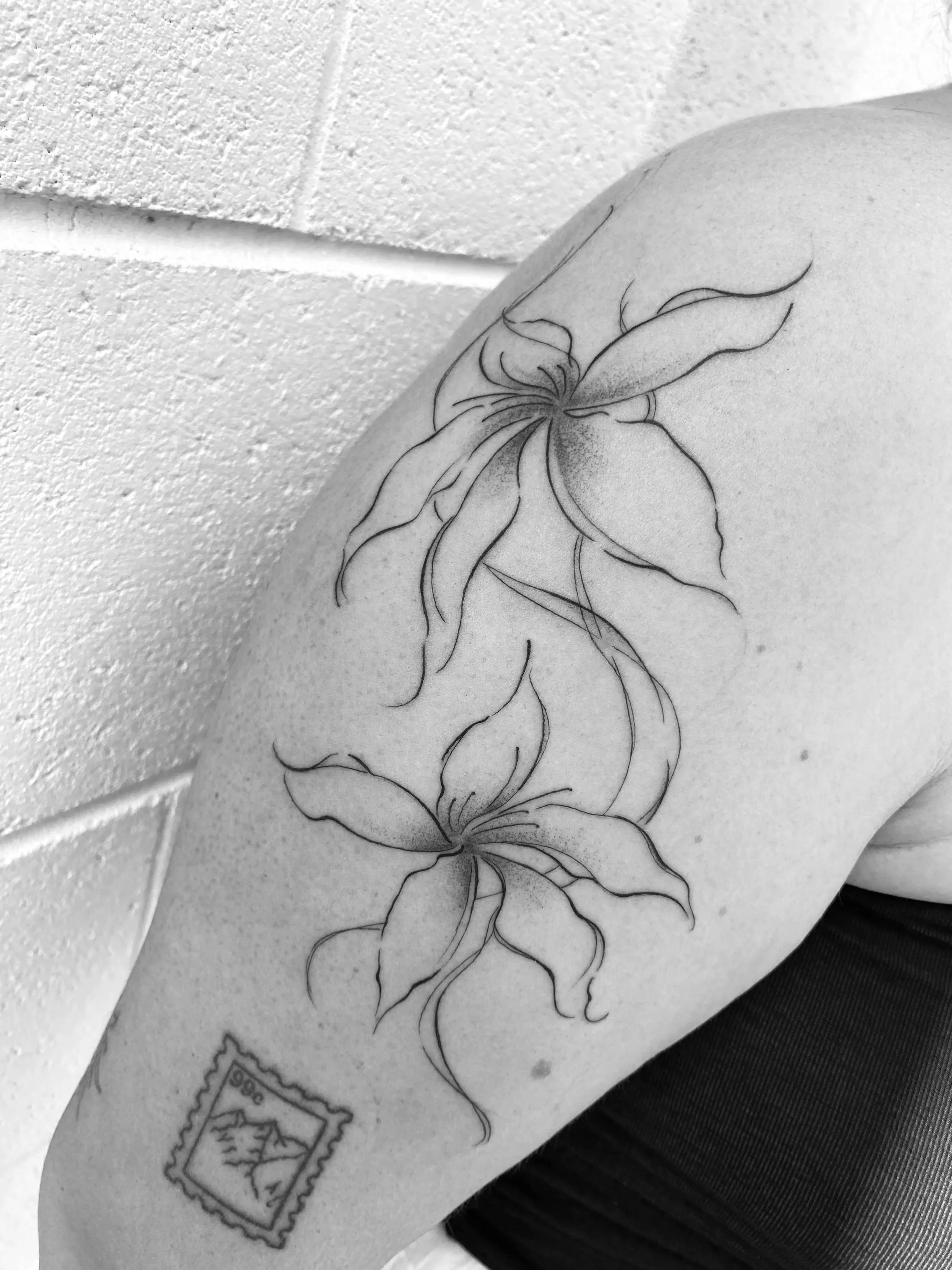 Black and white tattoo of two lilies on a person's upper arm, with a postage stamp featuring mountains and a mountain range at the bottom.