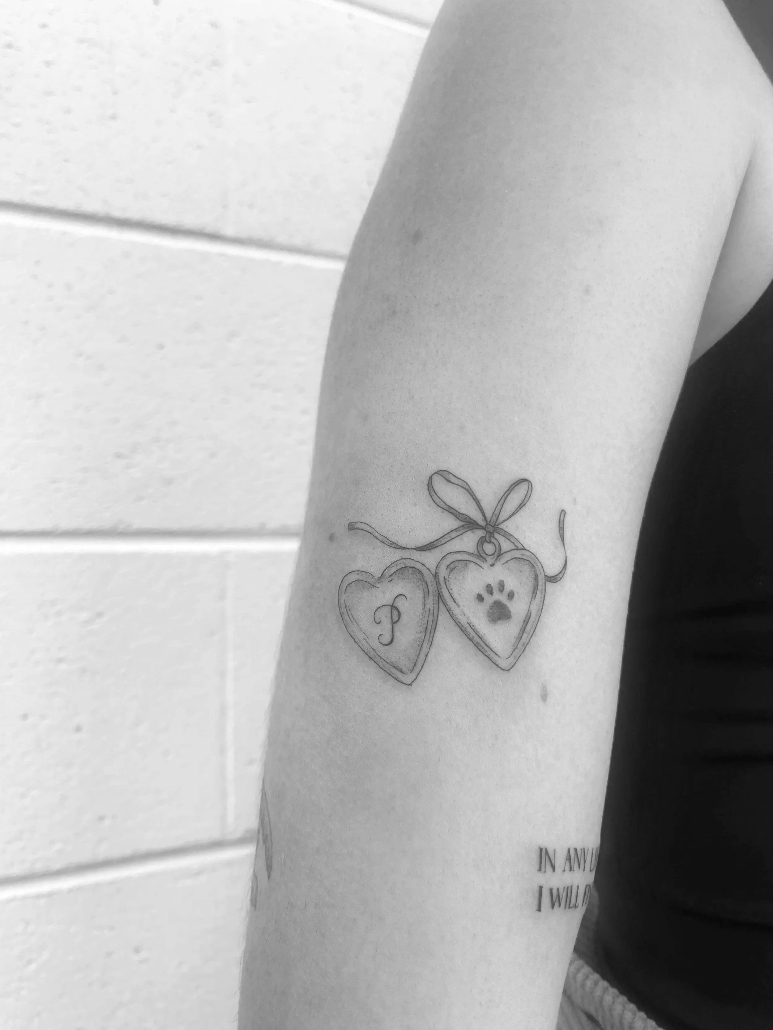 Tattoo of two heart-shaped charms, one with the letter 'P' and the other with a paw print, connected by a ribbon.
