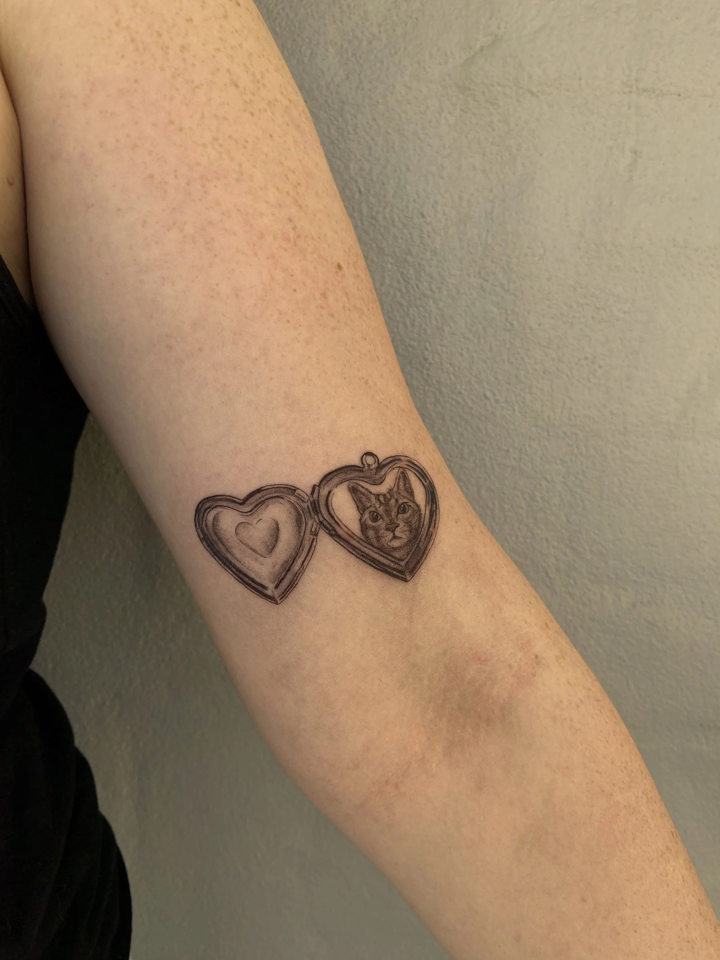 Tattoo of a heart-shaped locket with a cat's face inside, on a person's upper arm.