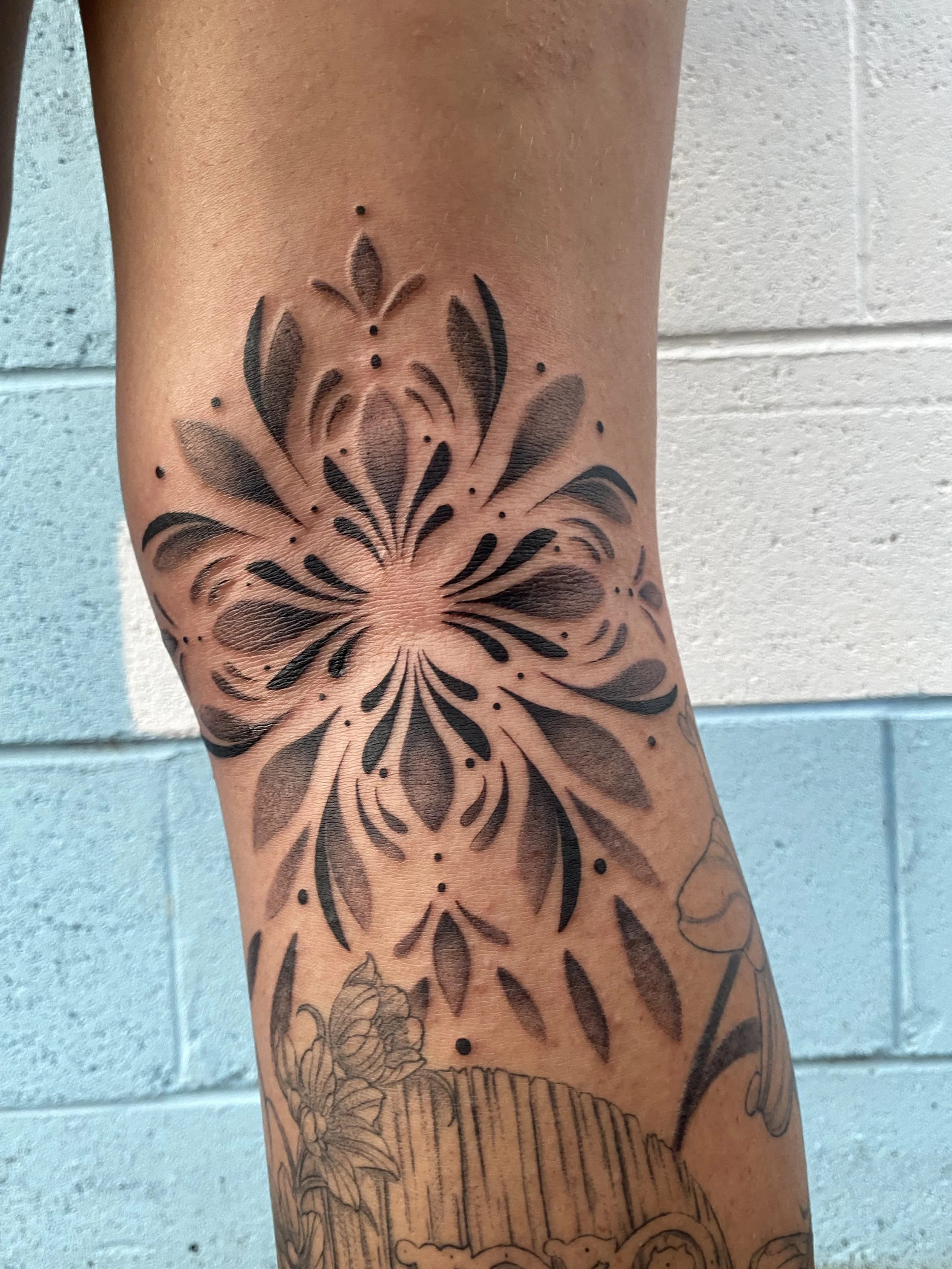 Black floral tattoo on person's forearm with detailed shading and abstract elements, against a light-colored brick wall.