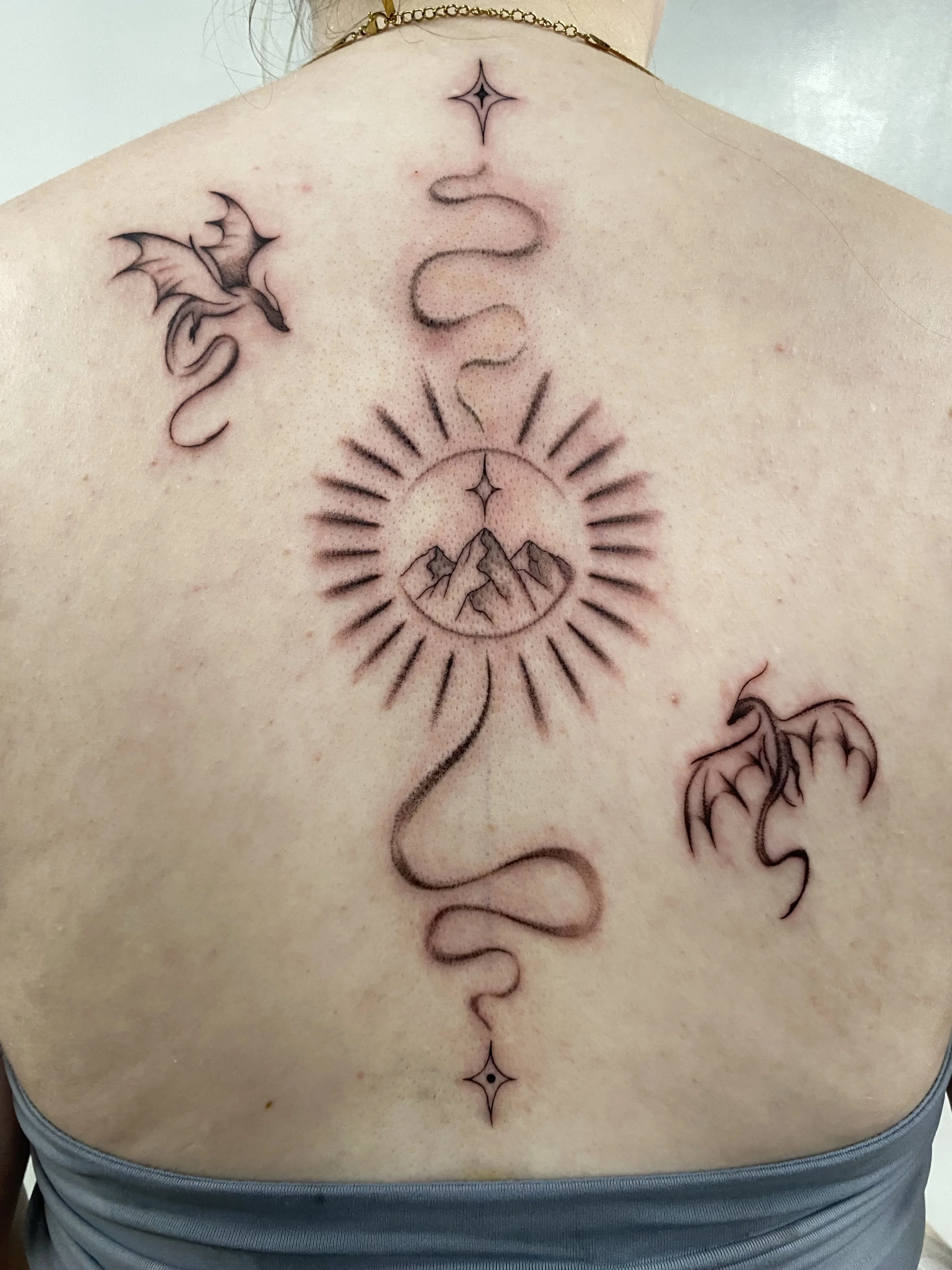Tattoo on person's back featuring a mountain scene inside a sun with rays, flanked by two dragons and decorative star shapes above and below.
