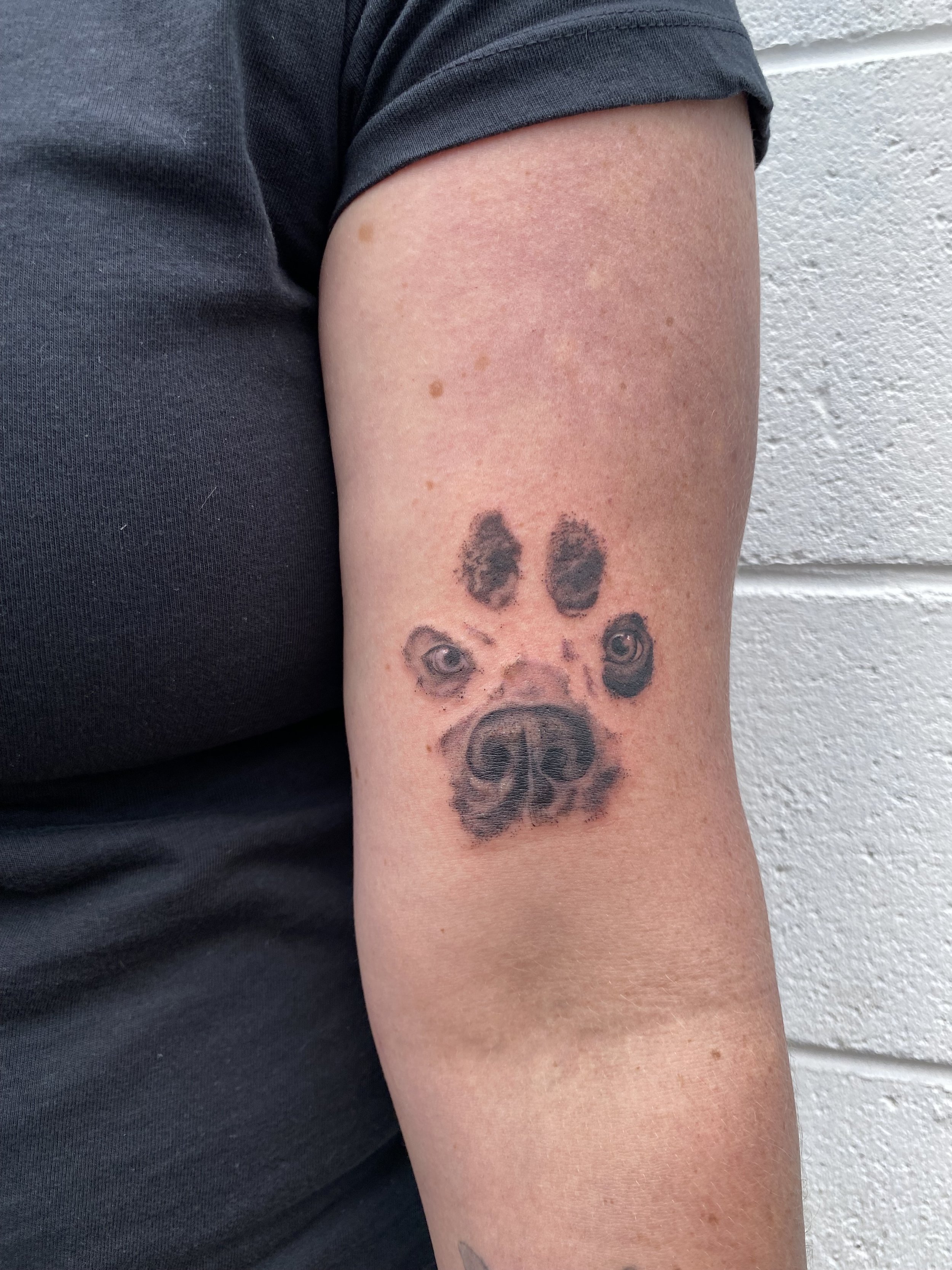 Tattoo of a dog's face on person's upper arm, with realistic eyes and nose, on light skin.