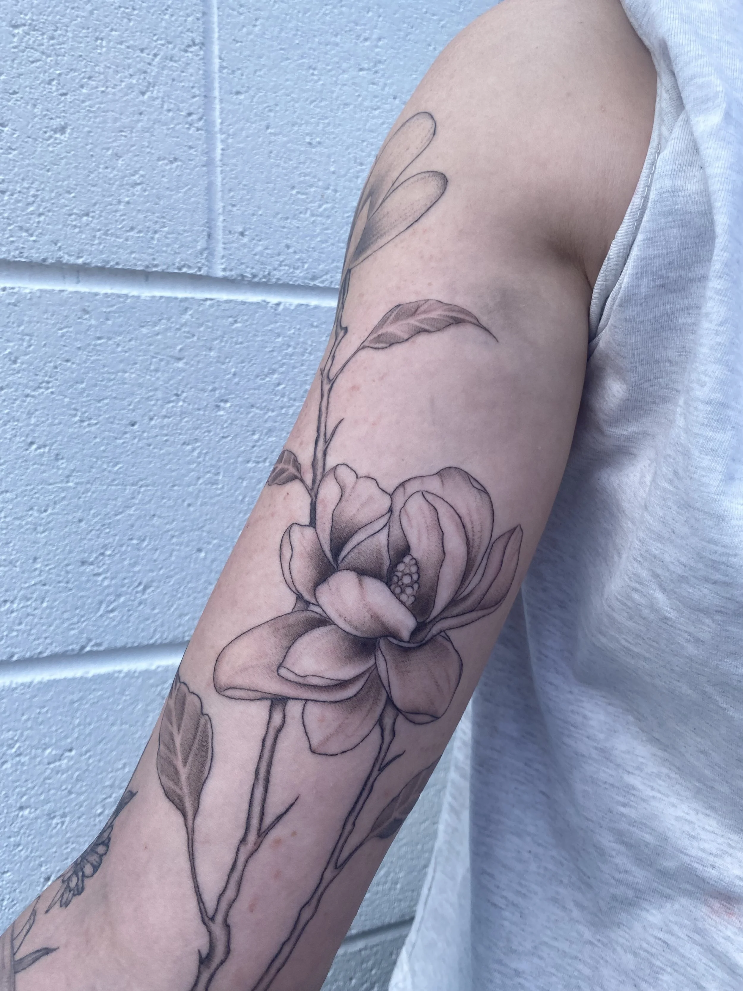 Black and gray floral tattoo on person's upper arm featuring a large blooming flower with detailed petals and leaves.