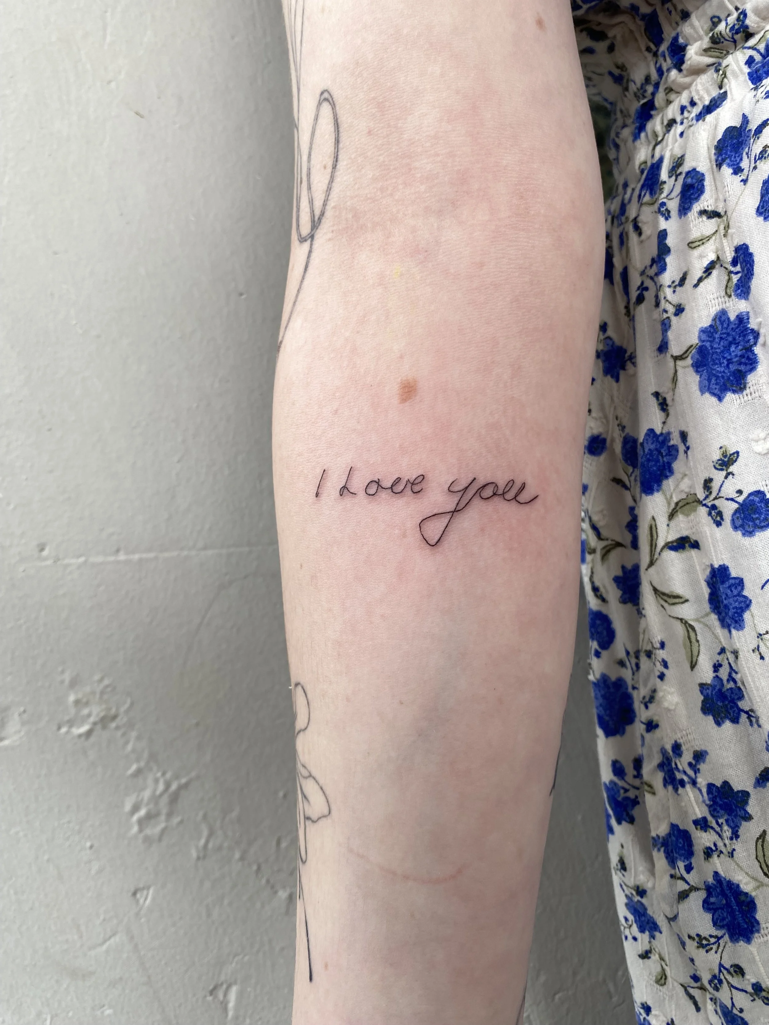 A tattoo of the words 'I love you' in cursive on a person's forearm, next to some line art of abstract shapes, with part of a blue floral dress visible in the background.